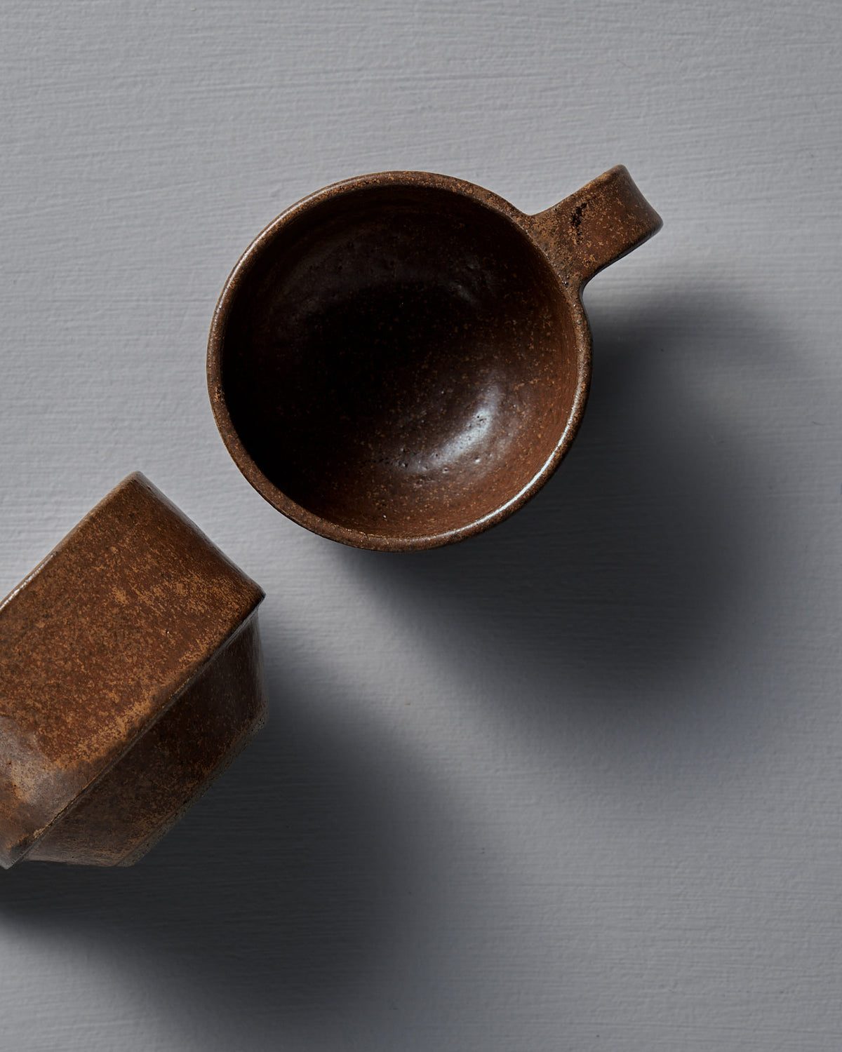 Two Studio Foon Everyday Stacker Cups – Small in Terracotta rest on a gray textured surface, one upright and the other tilted, casting soft shadows.