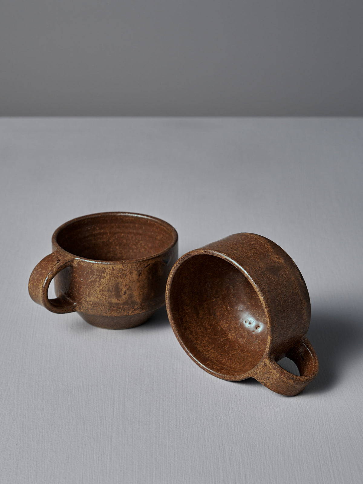 Two Everyday Stacker Cups – Small in Terracotta by Studio Foon, one upright and one on its side, rest on a light gray surface against a plain gray background.