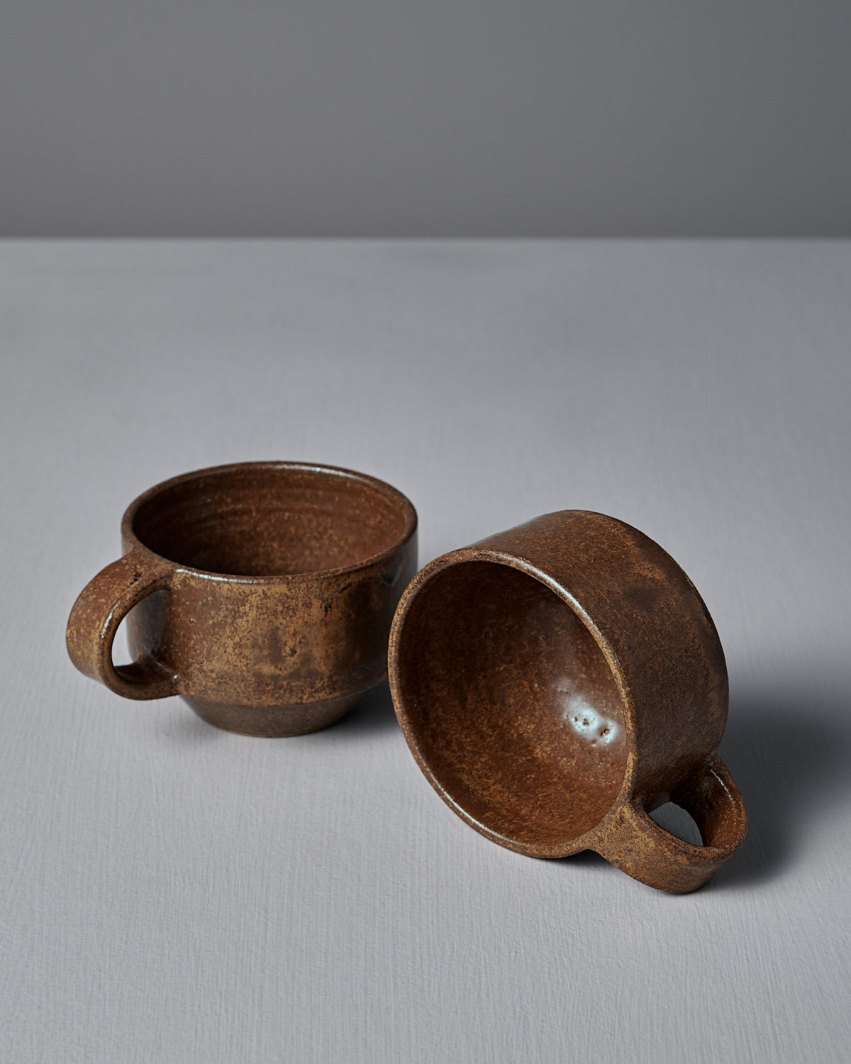 Two Everyday Stacker Cups – Small in Terracotta by Studio Foon, one upright and one on its side, rest on a light gray surface against a plain gray background.