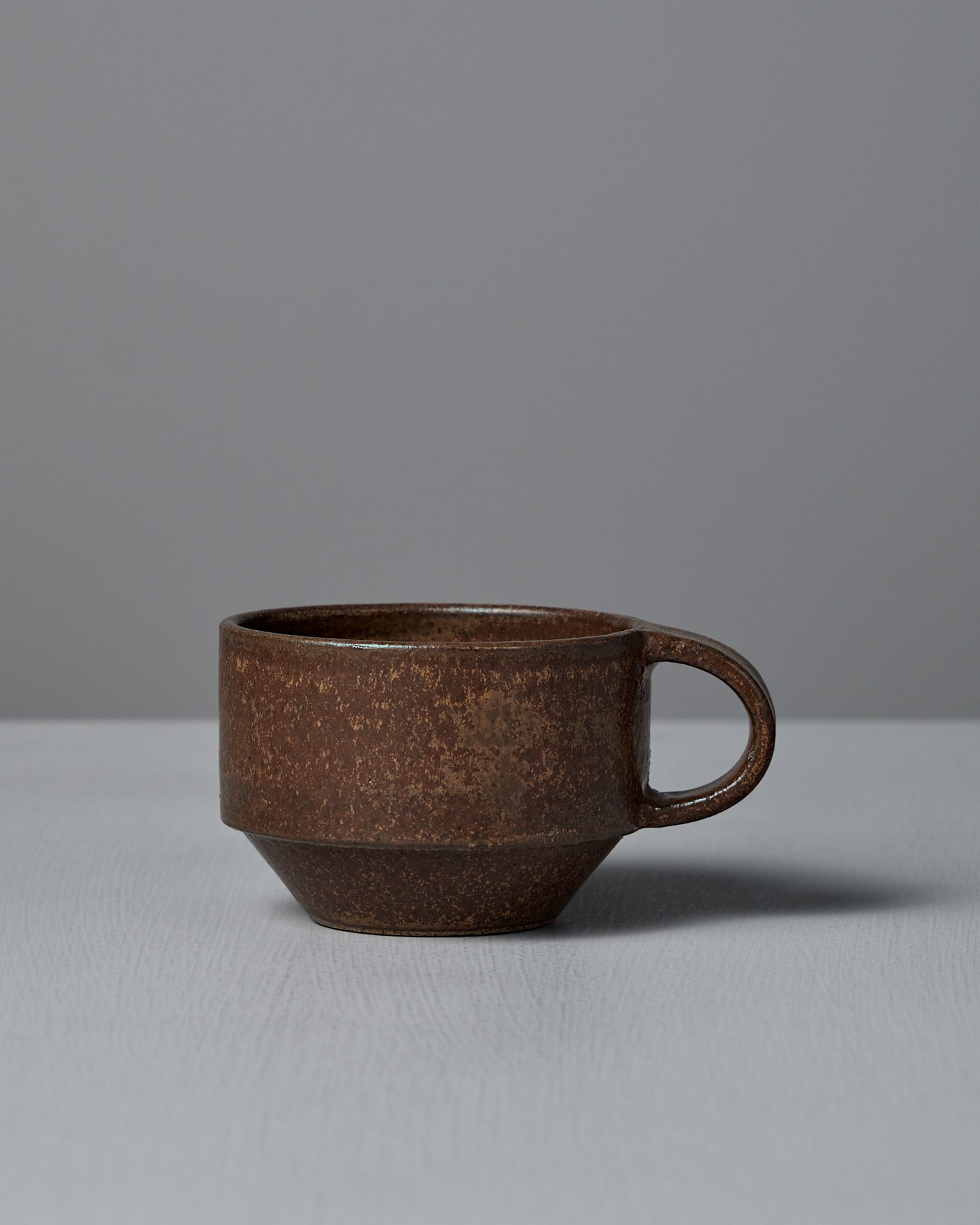 The Studio Foon Everyday Stacker Cup – Small in terracotta features a handle and sits on a light gray surface with a plain gray background, making it an ideal coffee cup for daily use.