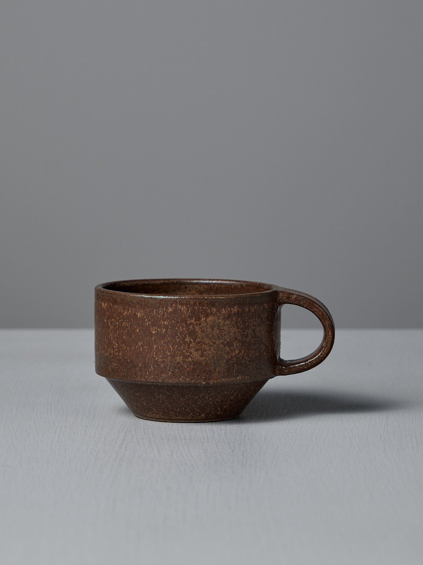 The Studio Foon Everyday Stacker Cup – Small in terracotta features a handle and sits on a light gray surface with a plain gray background, making it an ideal coffee cup for daily use.