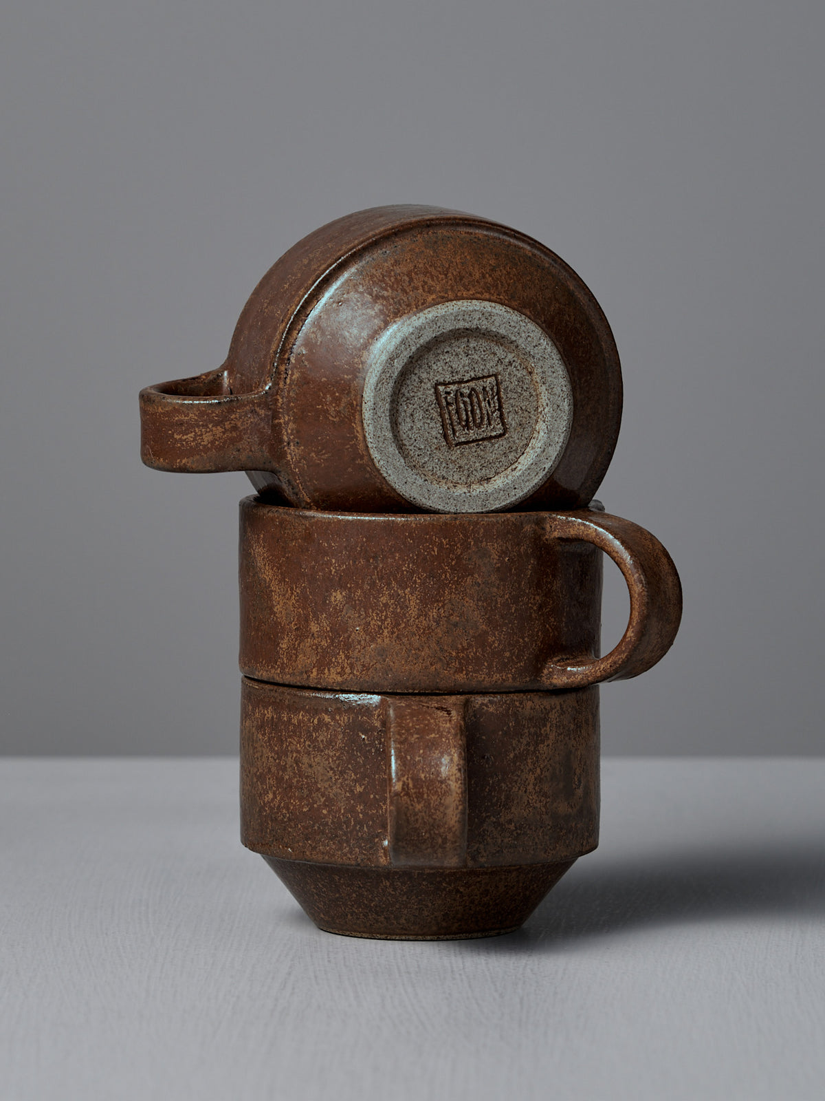Three Studio Foon Everyday Stacker Cups – Small in terracotta are stacked on a light grey surface, with the top cup turned sideways to display its bottom stamp.