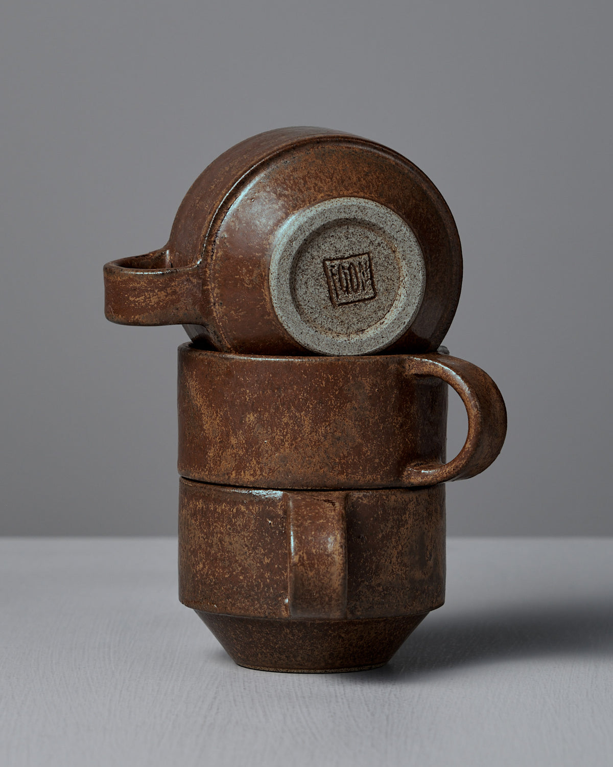 Three Studio Foon Everyday Stacker Cups – Small in terracotta are stacked on a light grey surface, with the top cup turned sideways to display its bottom stamp.