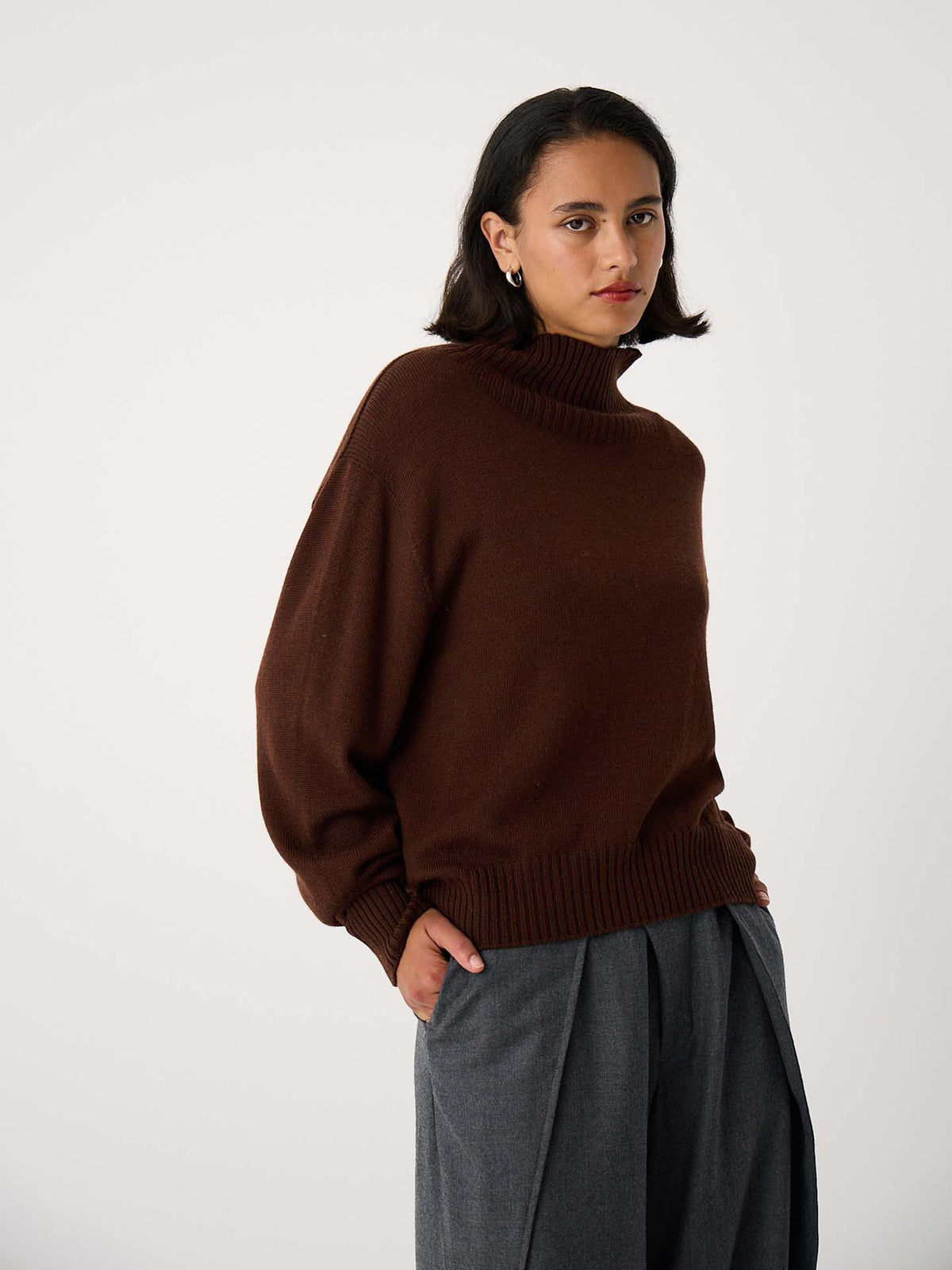 A person wears the Albers Turtle Neck – Chestnut, a relaxed fit brown turtleneck sweater, paired with gray pants and stands against a plain light background with hands in pockets.