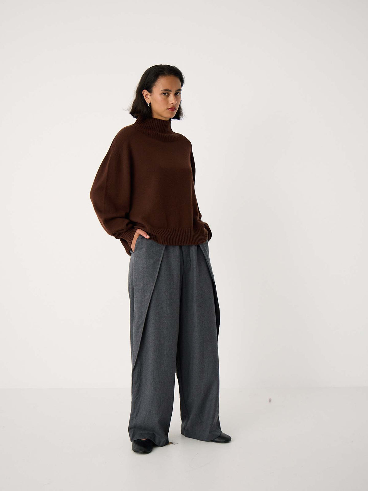 A person stands against a plain white background, wearing the Albers Turtle Neck – Chestnut and wide-leg gray trousers with hands in pockets.