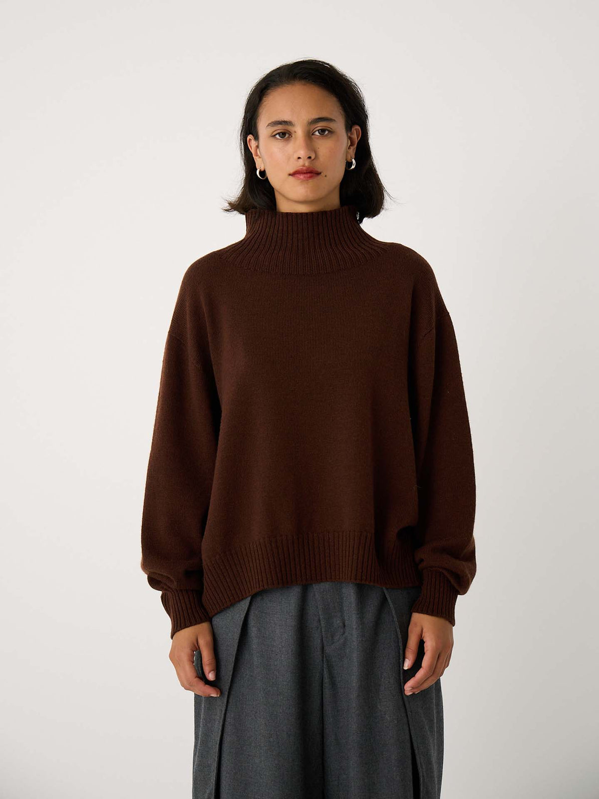 A person stands facing forward, wearing the Albers Turtle Neck – Chestnut sweater and gray wide-leg pants against a plain light background.