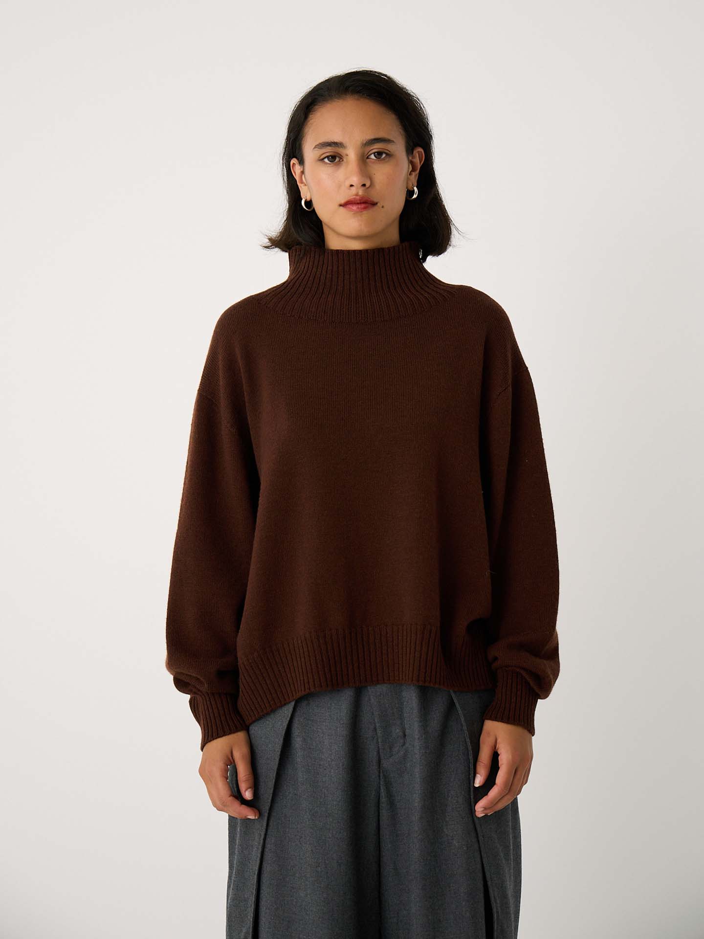 A person stands facing forward, wearing the Albers Turtle Neck – Chestnut sweater and gray wide-leg pants against a plain light background.
