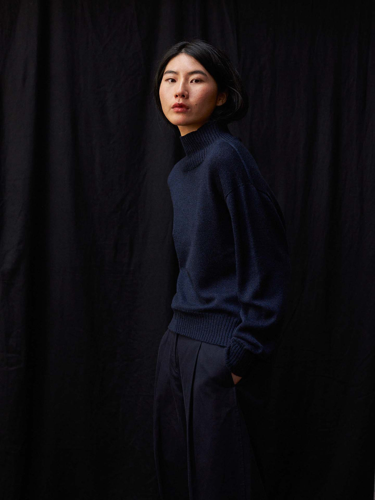A person stands before a dark backdrop, hands in pockets and neutral-faced, showcasing the relaxed fit and effortless style of the Albers Knit – Ink sweater paired with dark pants.
