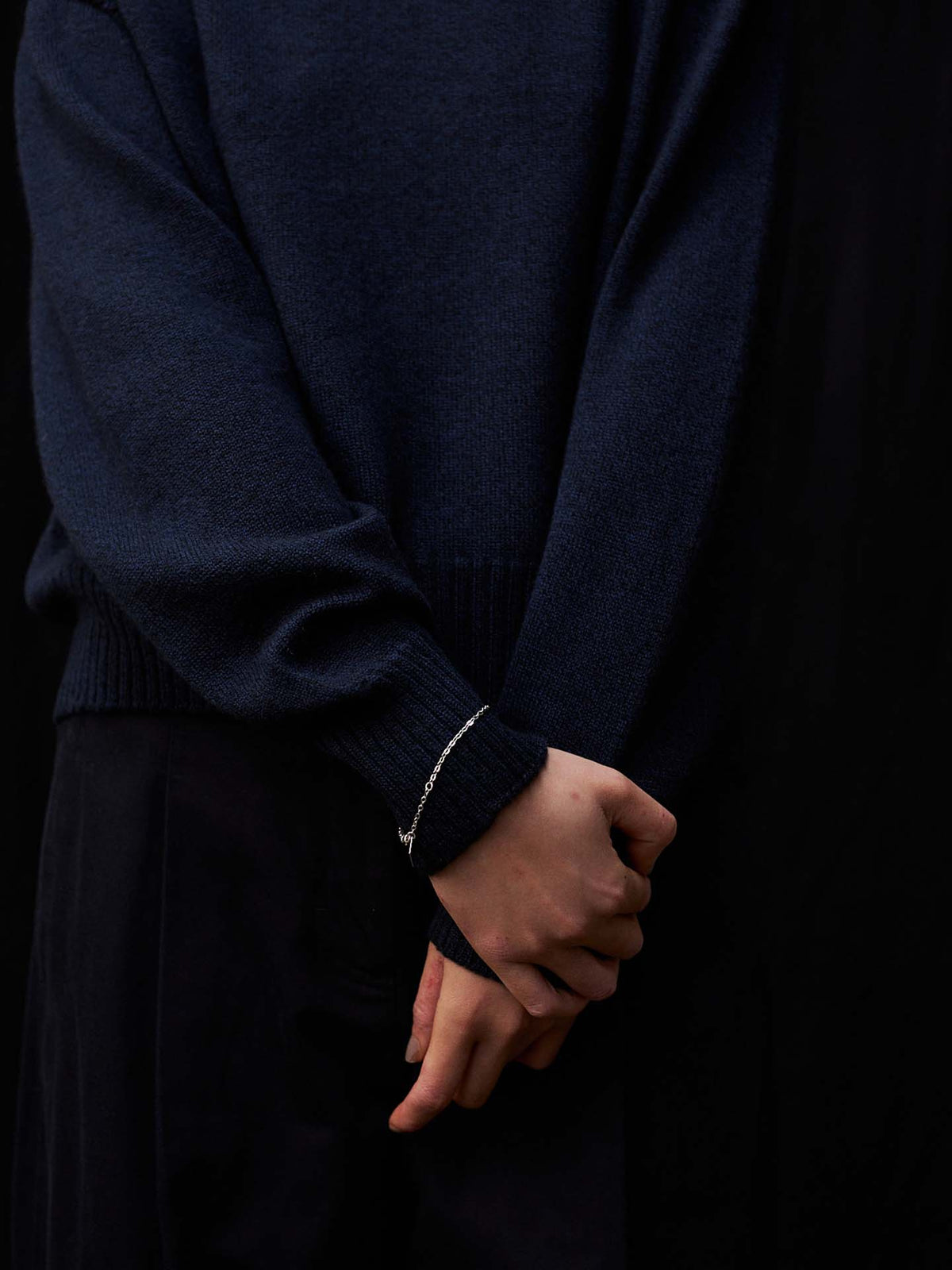 A person stands against a black background in the Albers Knit – Ink, a relaxed fit navy blue sweater, paired with dark pants and a thin bracelet on their left wrist, hands clasped behind their back.