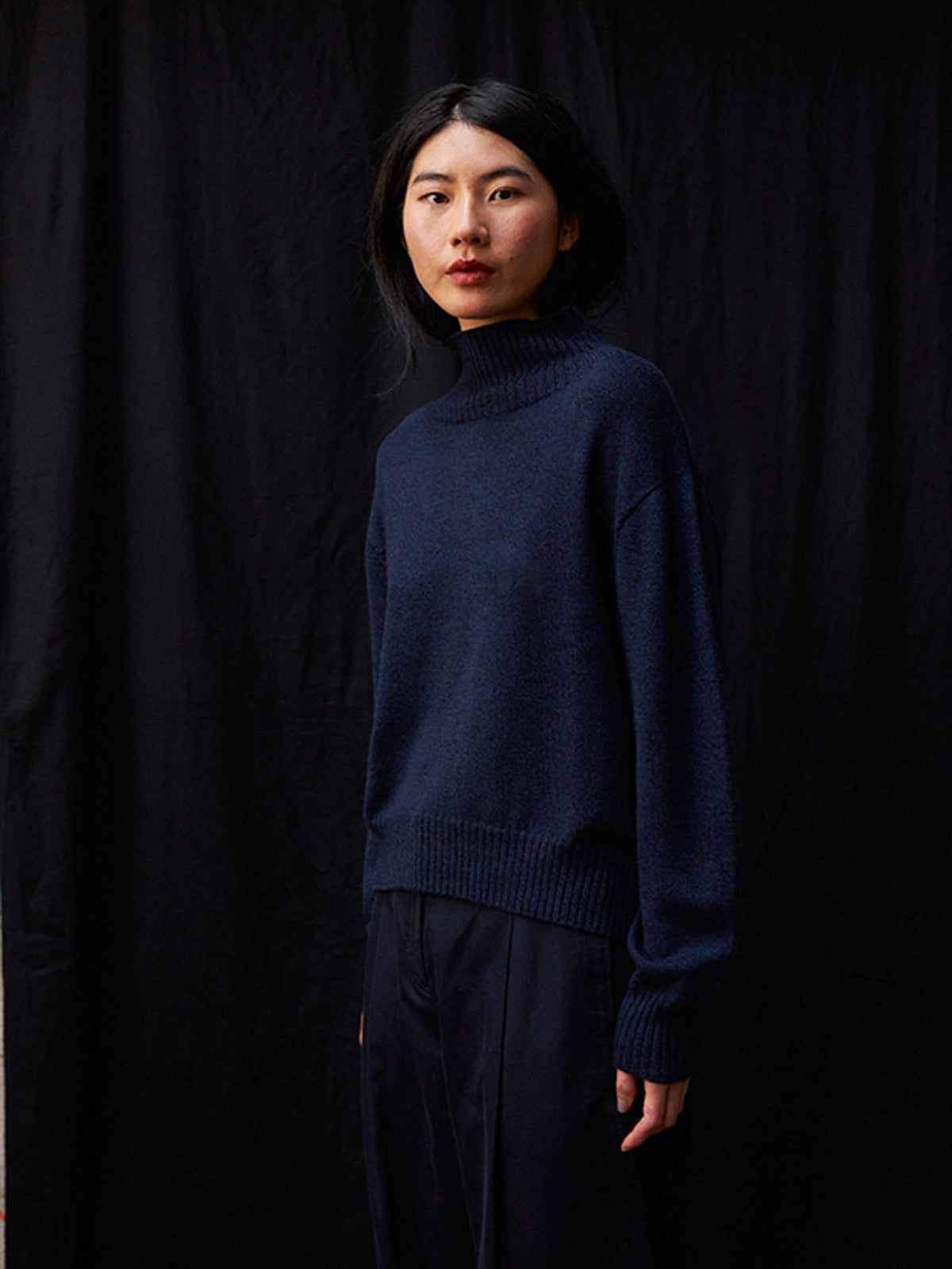 A person faces the camera with a neutral expression, dressed in the Albers Knit – Ink oversized turtleneck sweater and dark pants against a dark backdrop.
