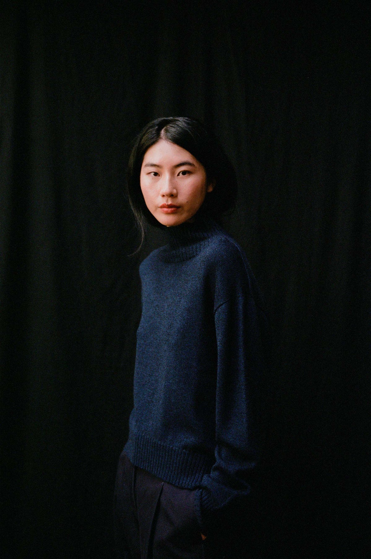 A person with long dark hair wears the Albers Knit – Ink sweater in a relaxed fit, standing before a black background and looking at the camera with a neutral expression.