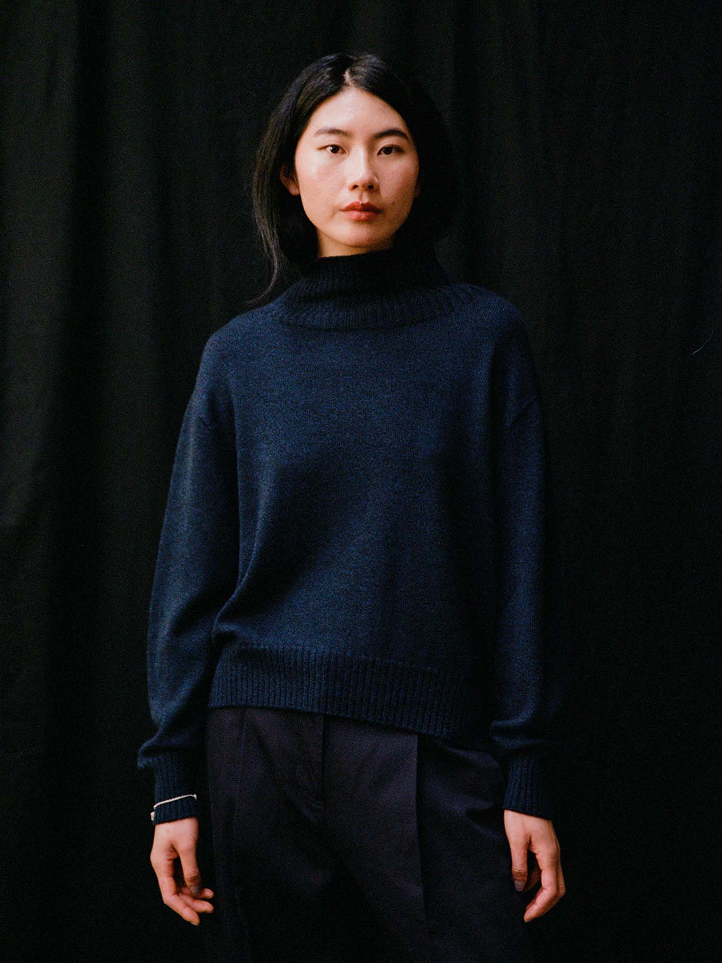 A person with long dark hair, dressed in an Albers Knit – Ink navy blue relaxed fit sweater and dark pants, stands against a black background.