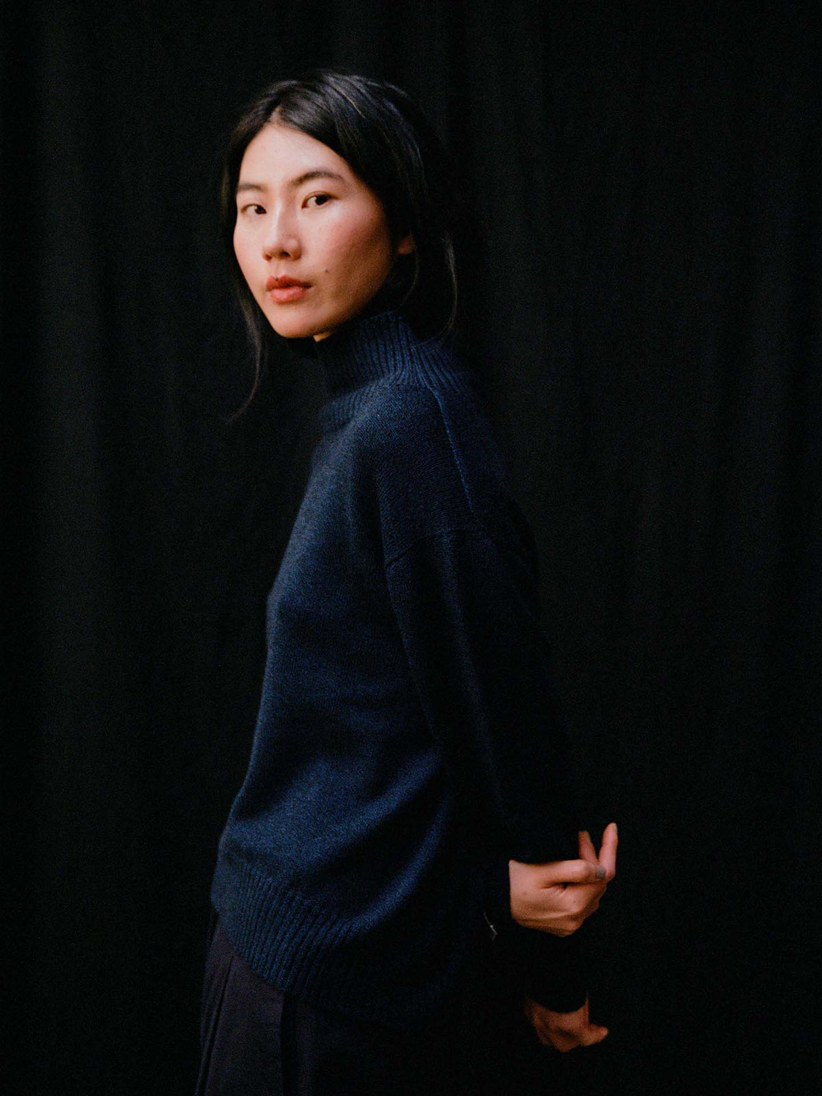 A person with long dark hair, wearing the Albers Knit – Ink, stands against a black background, turned slightly to the side with hands behind their back.