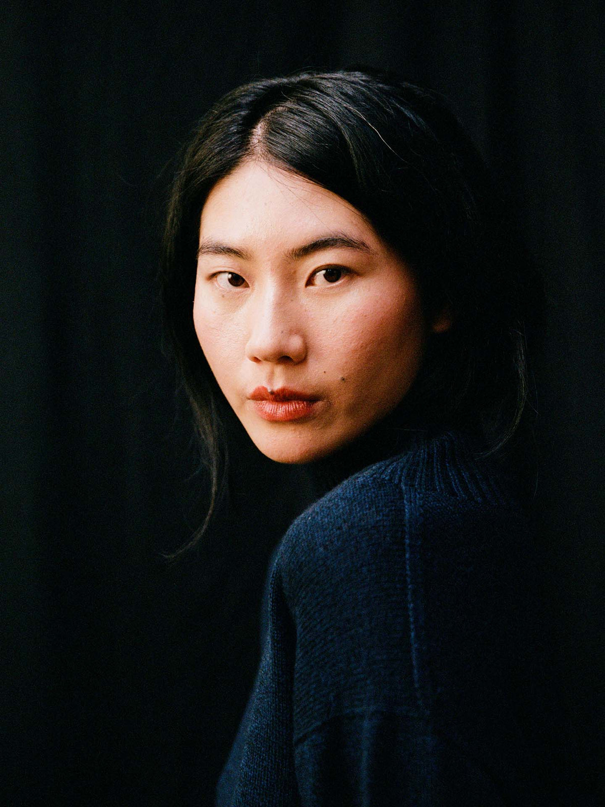 A person with long black hair wears the Albers Knit – Ink, an oversized turtleneck sweater, and looks directly at the camera against a black background.