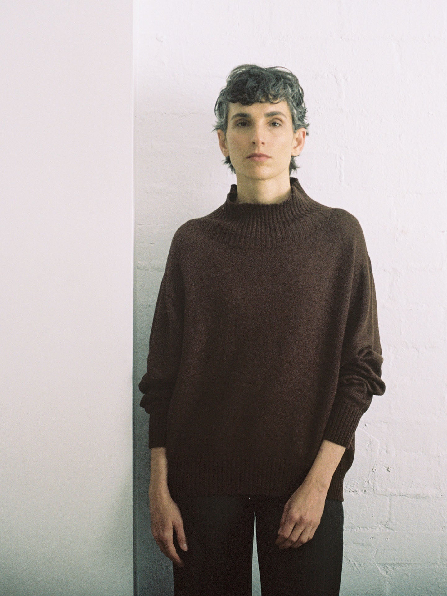 A relaxed person with short, curly hair stands against a white wall, wearing the oversized Albers Turtle Neck in Chestnut by Francie.