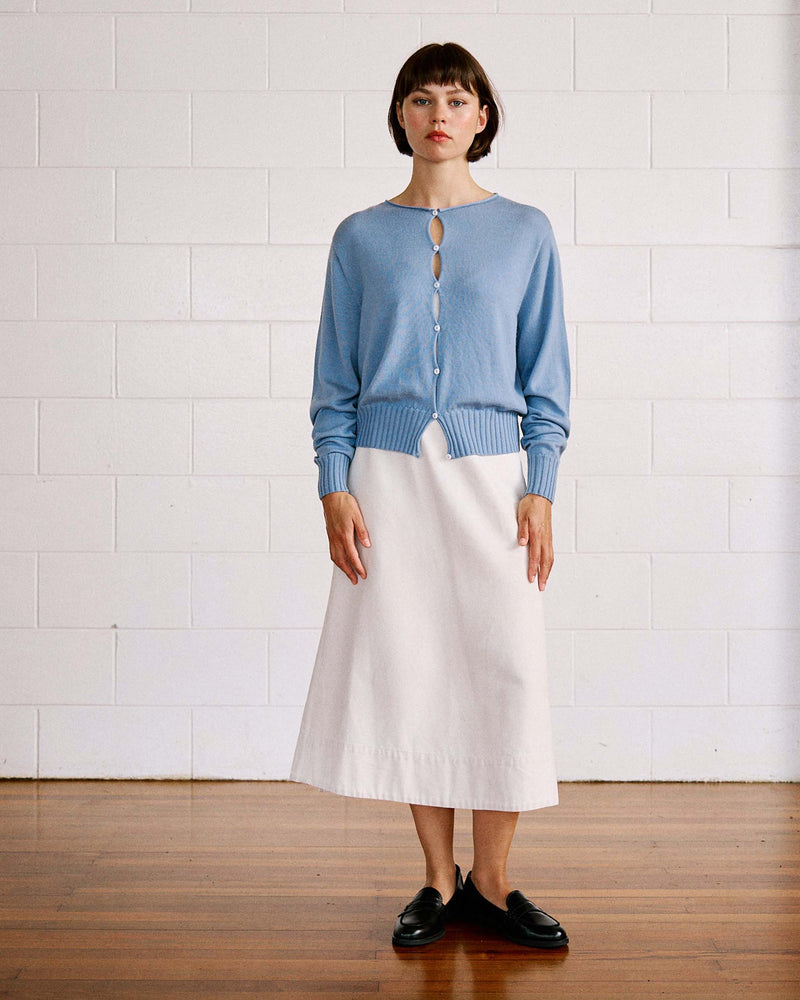 Woman standing indoors, wearing a light blue buttoned cardigan, white A-line skirt, and black loafers, against a white brick wall and wooden floor.