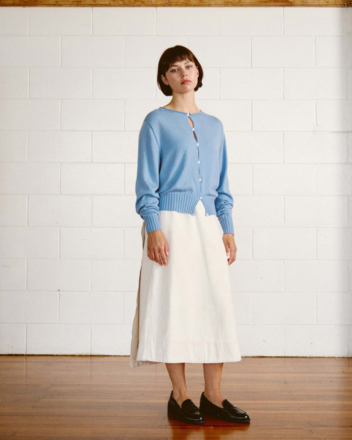 Person stands indoors against a white brick wall, wearing a light blue cardigan, white skirt, and black loafers on a wooden floor.