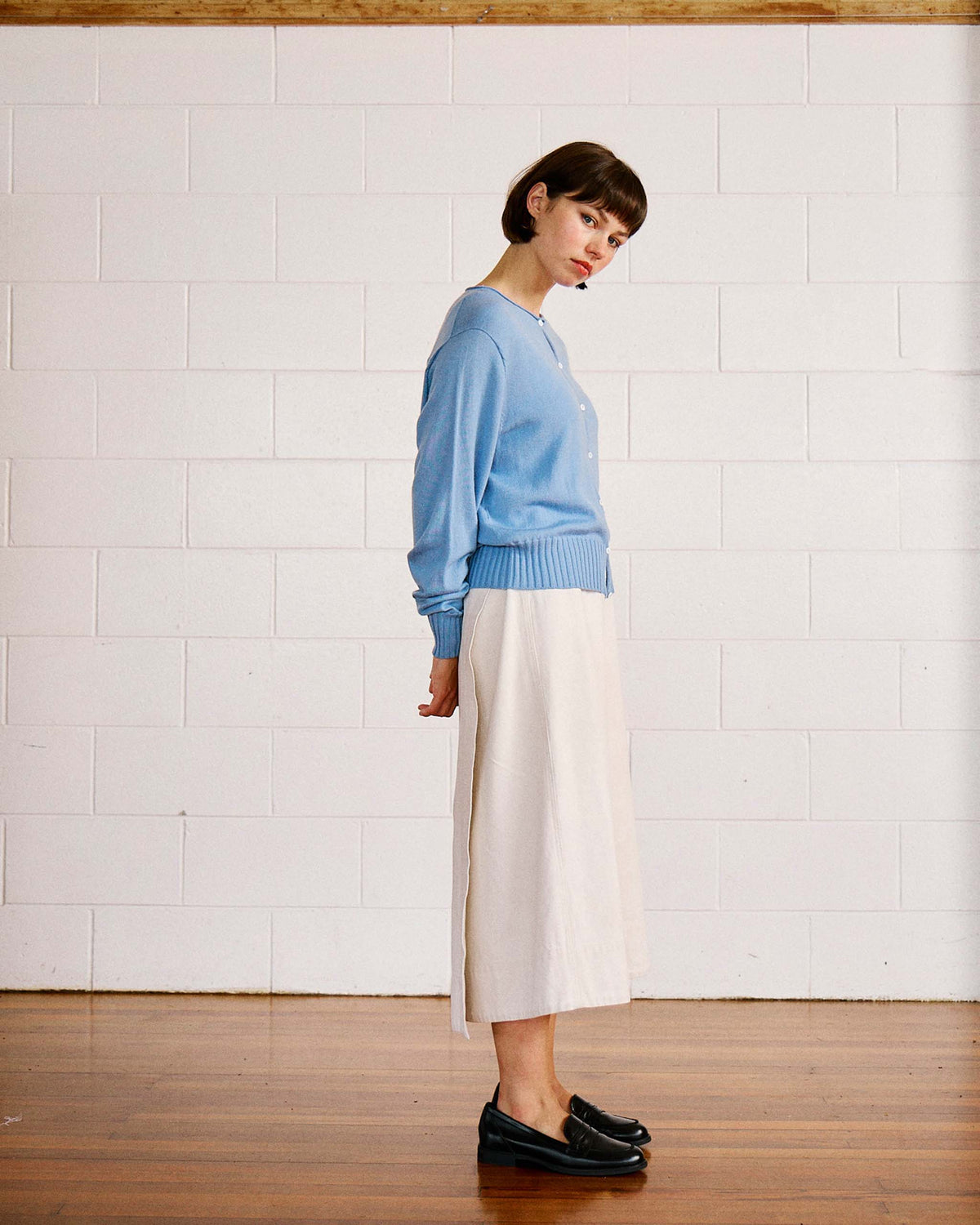 Woman standing sideways in front of a white brick wall, wearing a light blue sweater, cream midi skirt, and black loafers on a wooden floor.