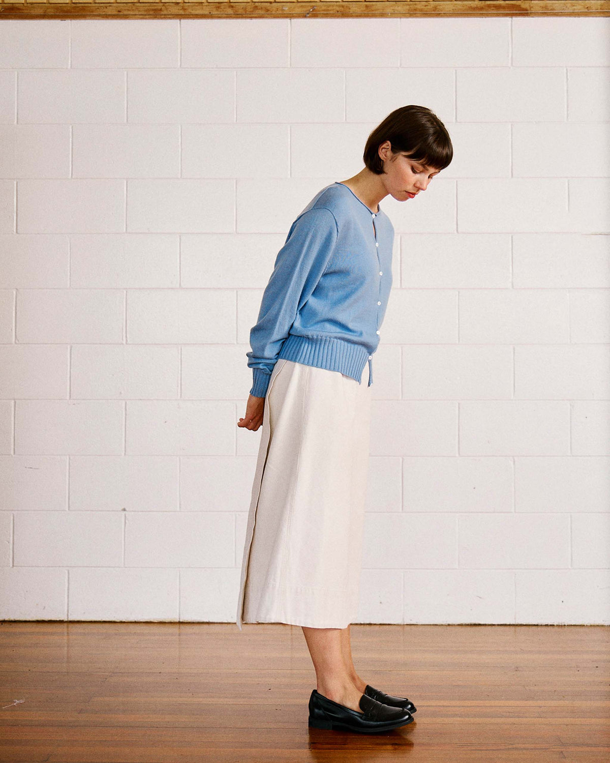 A woman stands sideways with her head down, wearing a blue sweater, white skirt, and black shoes, against a white brick wall and wooden floor.