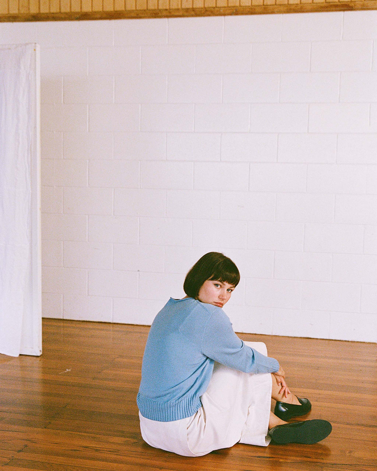 A woman with short hair, wearing a blue sweater and white skirt, sits on a wooden floor in front of a white brick wall, looking toward the camera.