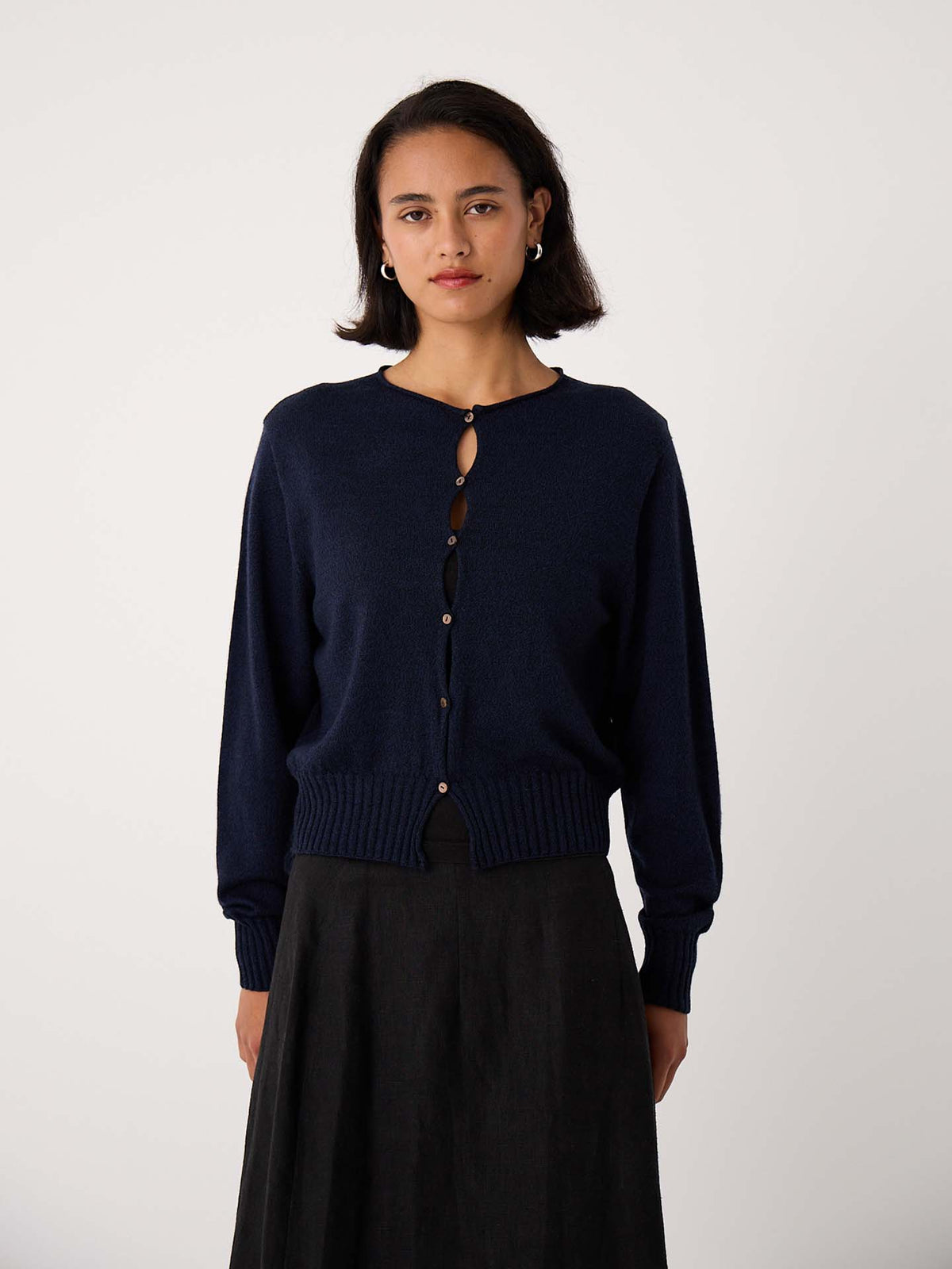 Petite woman stands against a plain background, facing the camera with a neutral expression, wearing the Anni Cardigan – Ink in a relaxed fit along with a dark skirt.