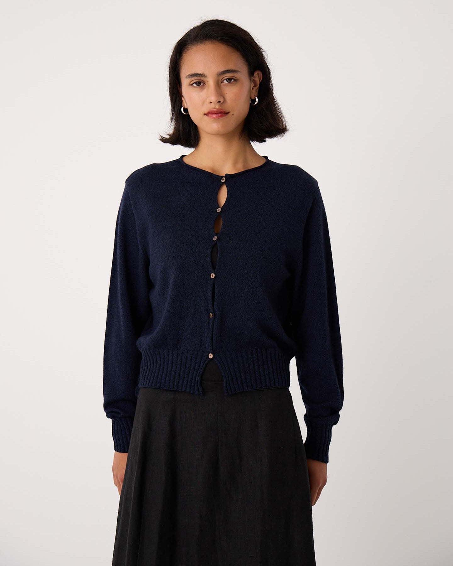 Petite woman stands against a plain background, facing the camera with a neutral expression, wearing the Anni Cardigan – Ink in a relaxed fit along with a dark skirt.