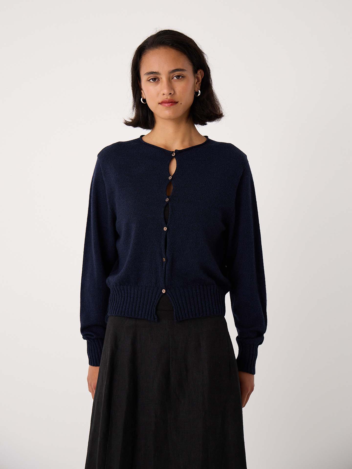 Petite woman stands against a plain background, facing the camera with a neutral expression, wearing the Anni Cardigan – Ink in a relaxed fit along with a dark skirt.