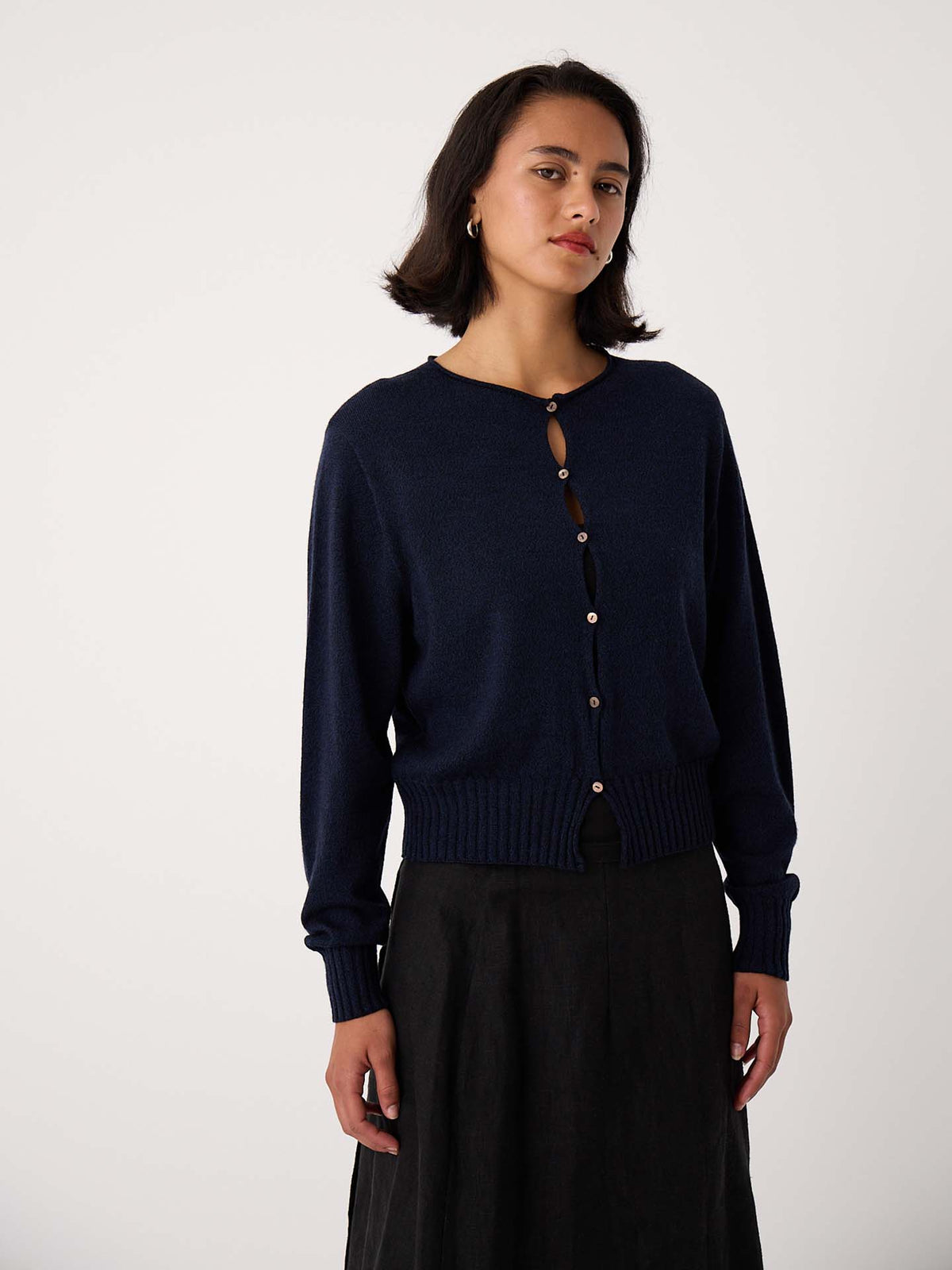 A petite woman wears the Anni Cardigan – Ink, a navy cropped button-up, paired with a black skirt, standing against a plain light background.