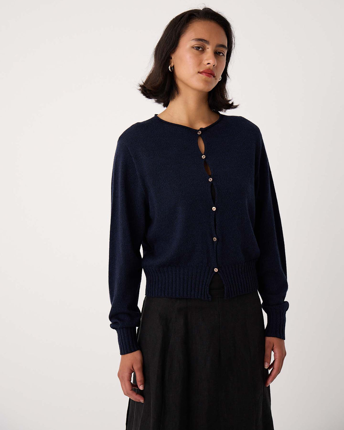 A petite woman wears the Anni Cardigan – Ink, a navy cropped button-up, paired with a black skirt, standing against a plain light background.