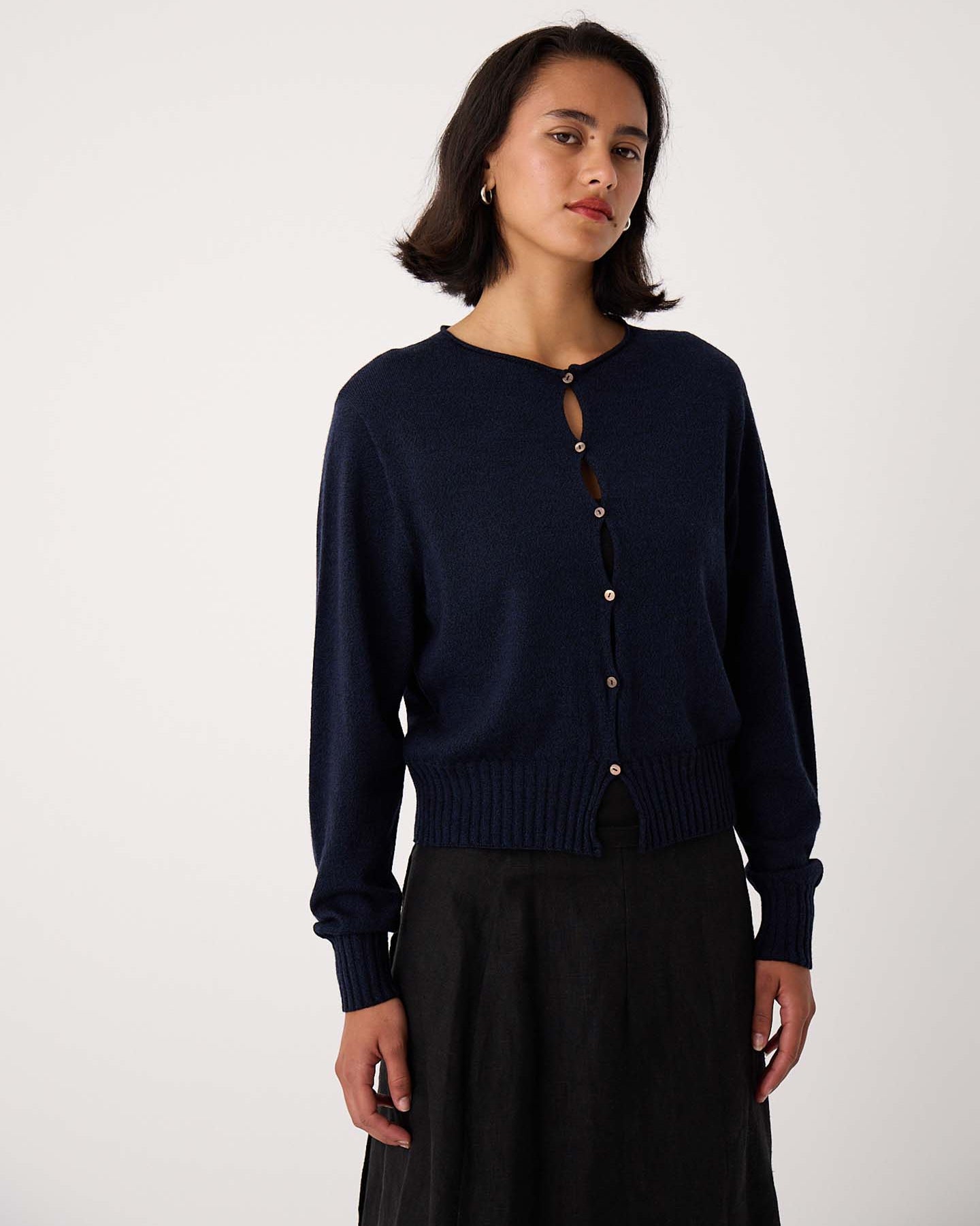 Petite woman stands against a plain background, facing the camera with a neutral expression, wearing the Anni Cardigan – Ink in a relaxed fit along with a dark skirt.