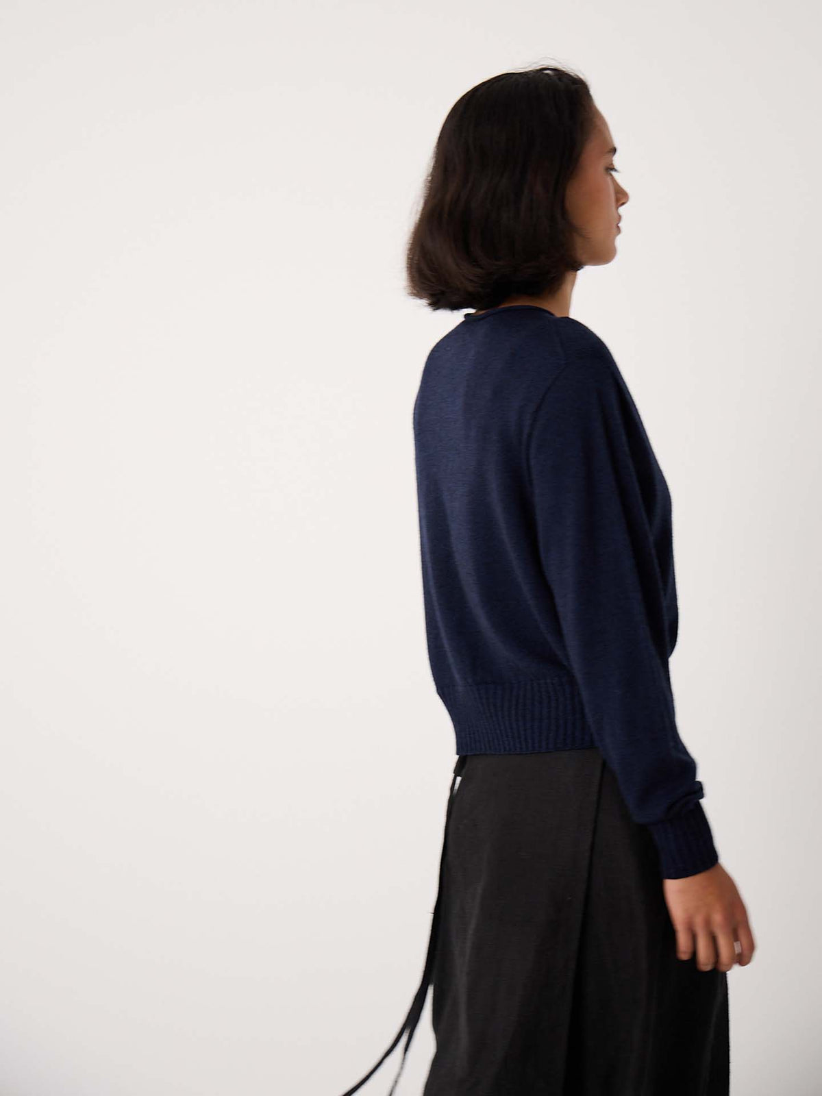 Petite person with short dark hair wears the Anni Cardigan in Ink, a relaxed fit navy blue sweater, paired with a black skirt, standing sideways against a plain light background.