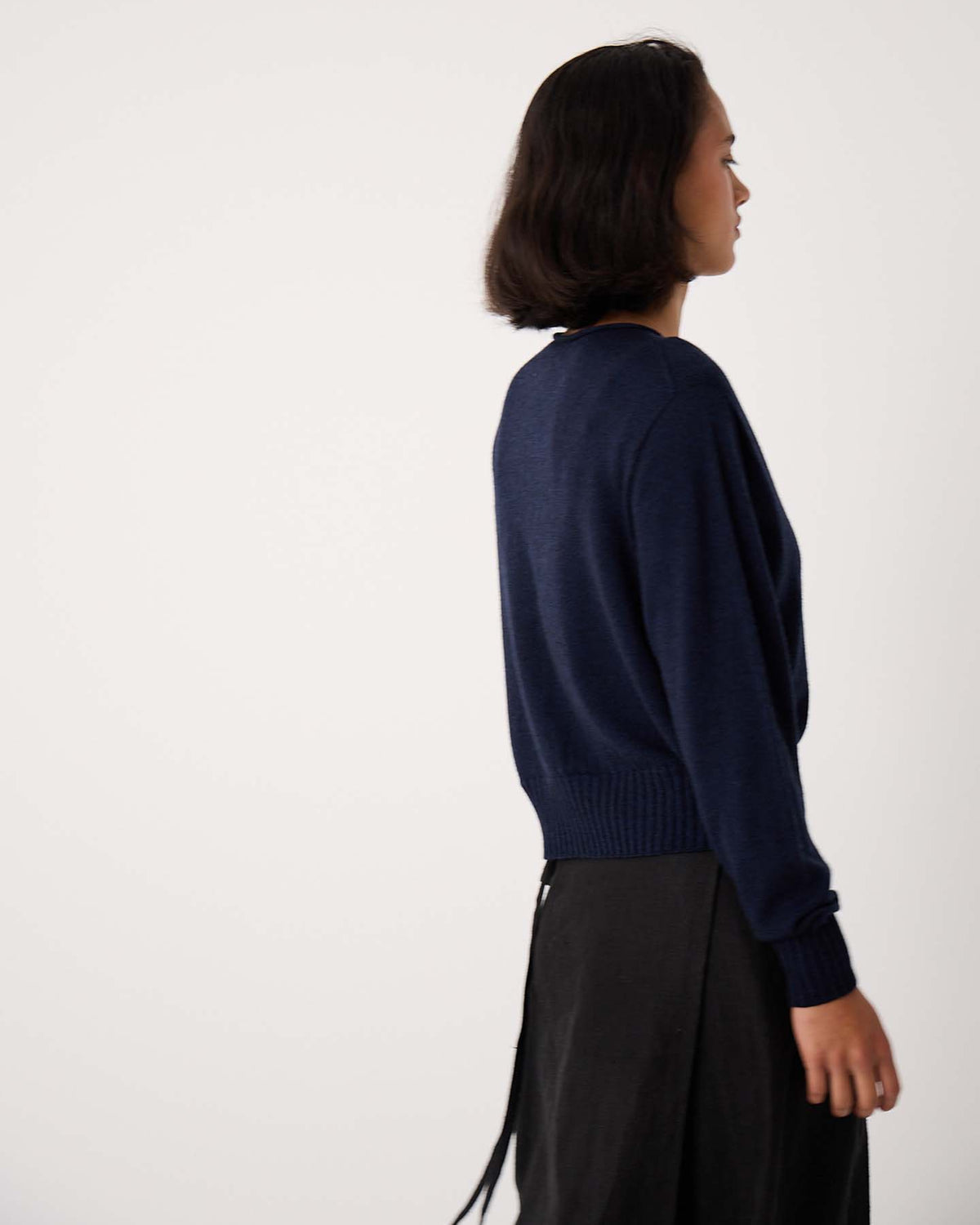 Petite person with short dark hair wears the Anni Cardigan in Ink, a relaxed fit navy blue sweater, paired with a black skirt, standing sideways against a plain light background.