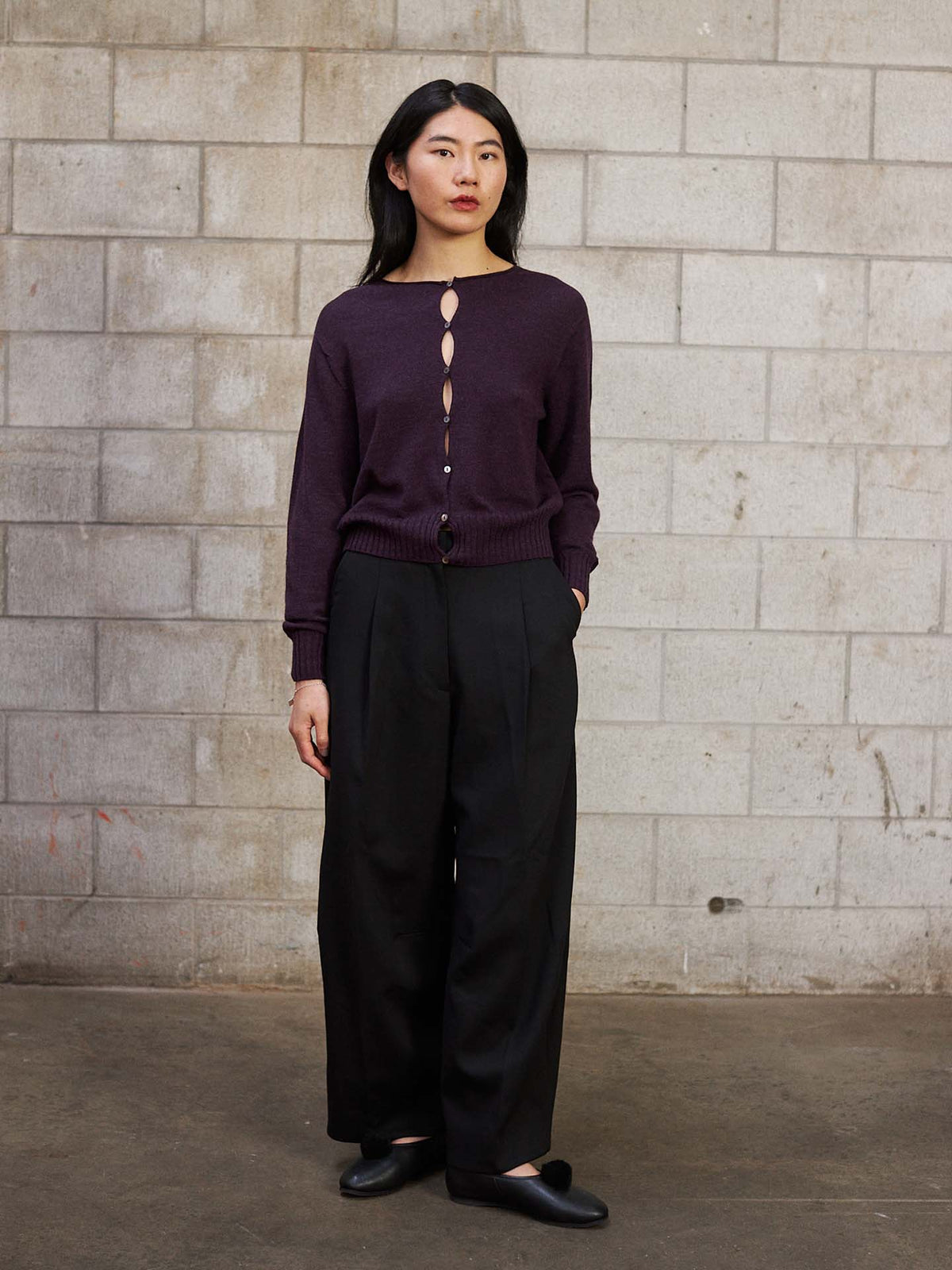 A woman stands against a cinder block wall, wearing the Anni Cardigan – Malbec in a relaxed fit, paired with black wide-leg pants and black shoes.