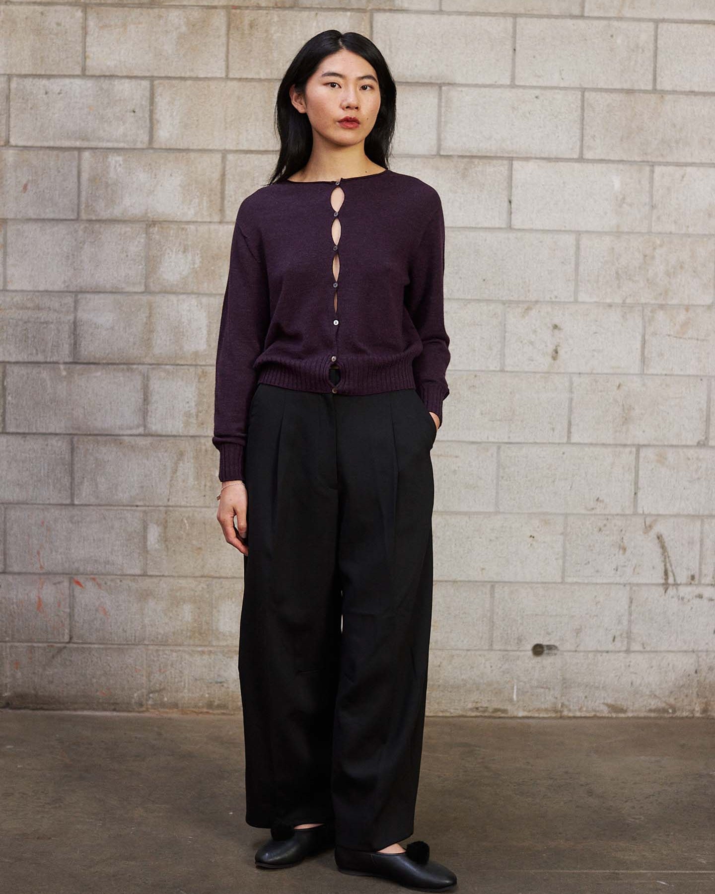 A woman stands against a cinder block wall, wearing the Anni Cardigan – Malbec in a relaxed fit, paired with black wide-leg pants and black shoes.