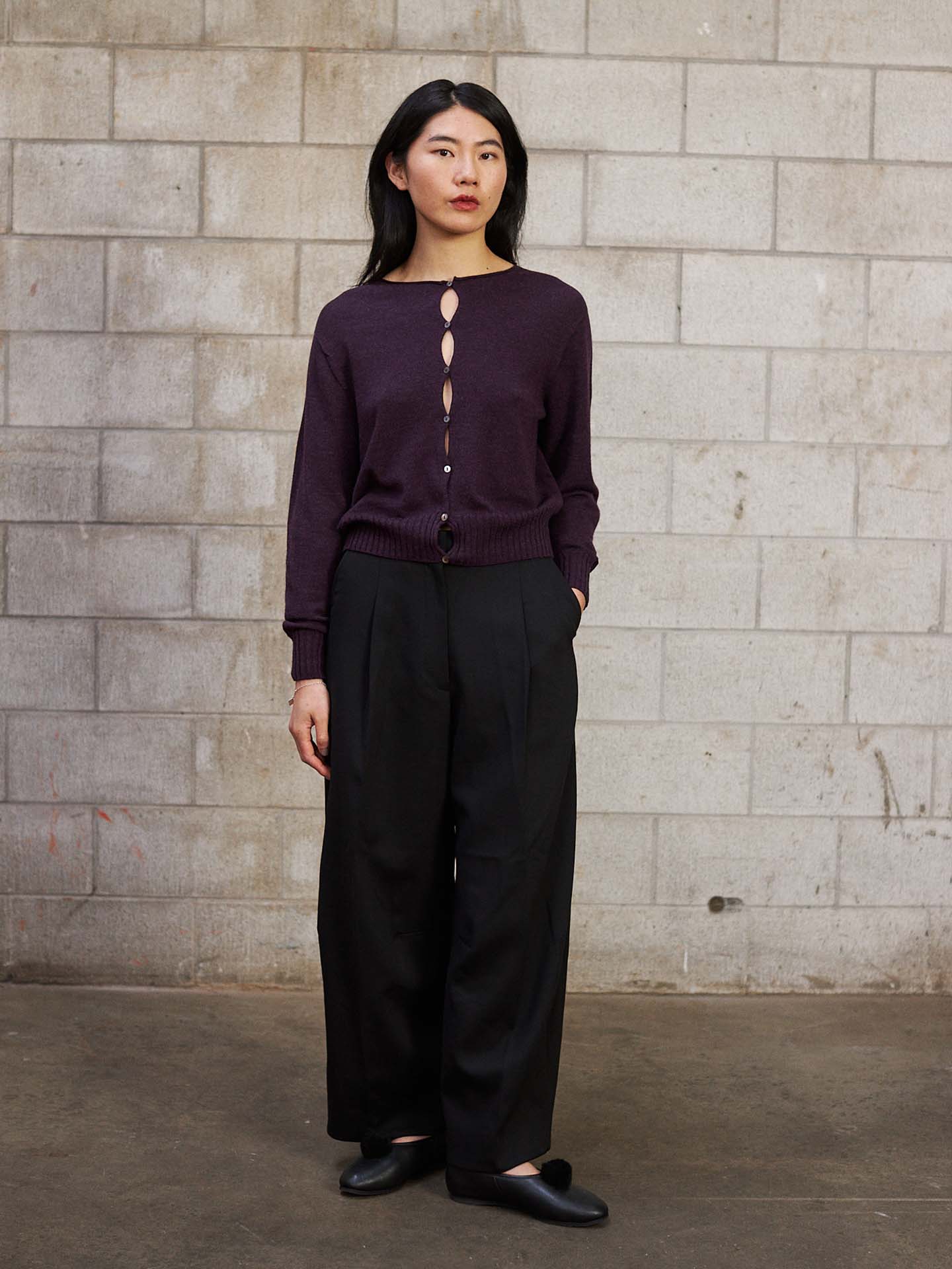 A woman stands against a cinder block wall, wearing the Anni Cardigan – Malbec in a relaxed fit, paired with black wide-leg pants and black shoes.