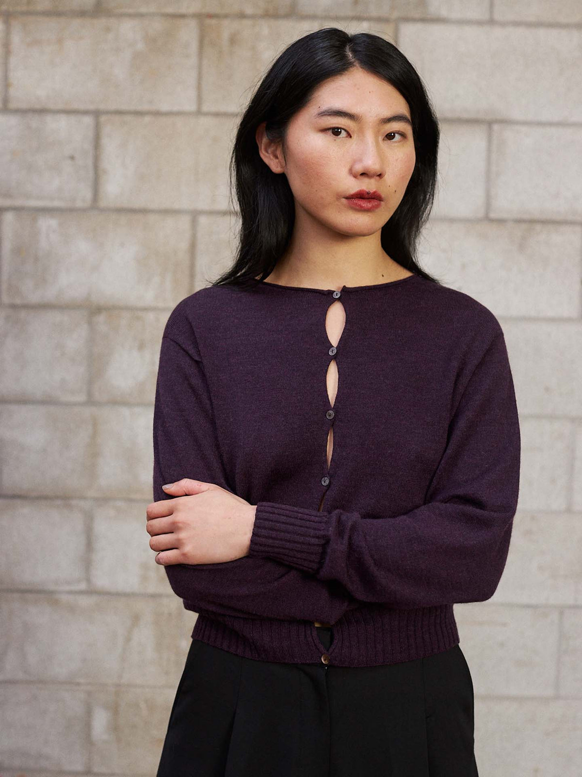 A person with long dark hair stands before a light brick wall, arms crossed, wearing the Anni Cardigan – Malbec in a relaxed fit with black pants.