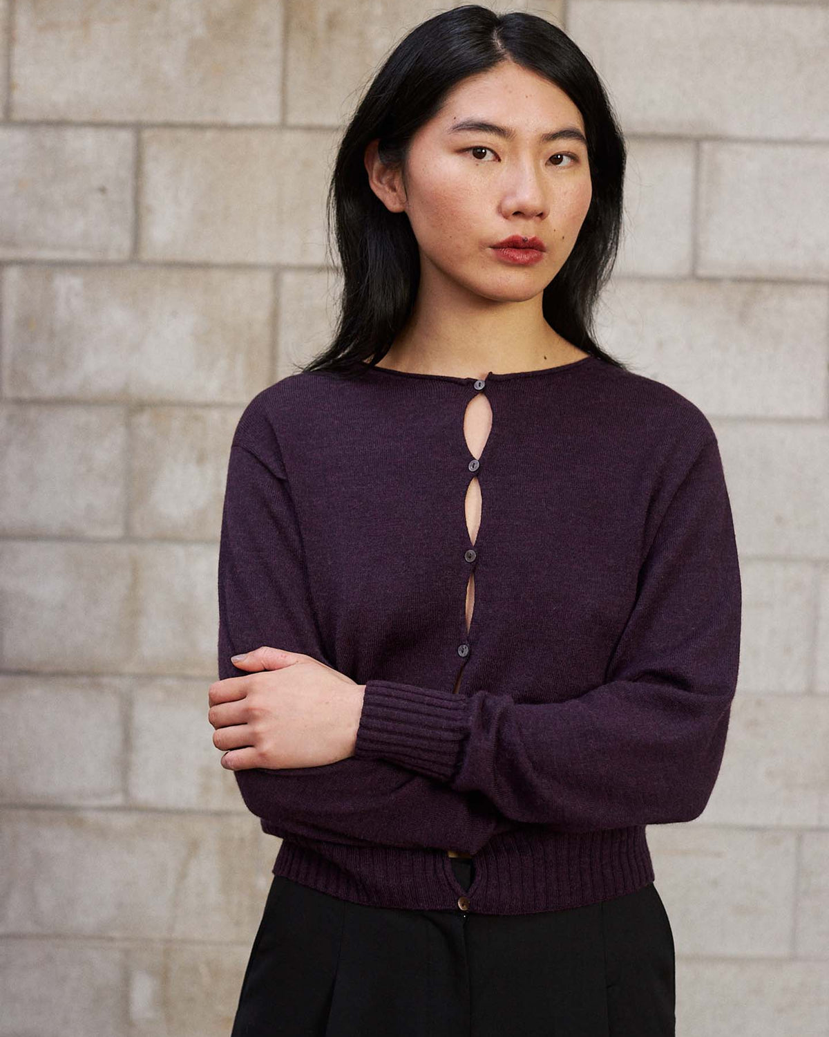A person with long dark hair stands before a light brick wall, arms crossed, wearing the Anni Cardigan – Malbec in a relaxed fit with black pants.