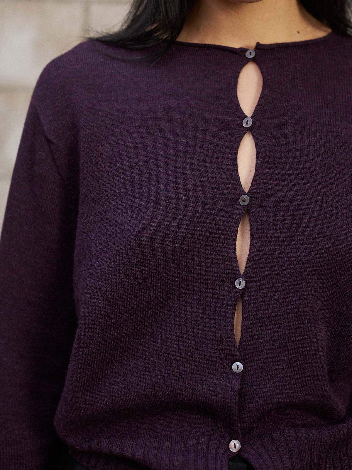Model wears the Anni Cardigan in Malbec—a dark purple sweater with an eye-catching button-up design featuring oval-shaped openings along the front and a relaxed fit for easy, effortless style.
