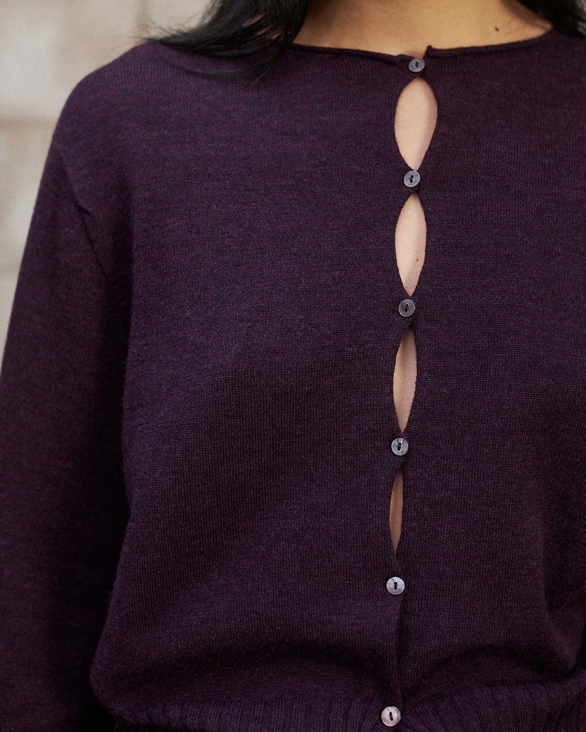 Model wears the Anni Cardigan in Malbec—a dark purple sweater with an eye-catching button-up design featuring oval-shaped openings along the front and a relaxed fit for easy, effortless style.