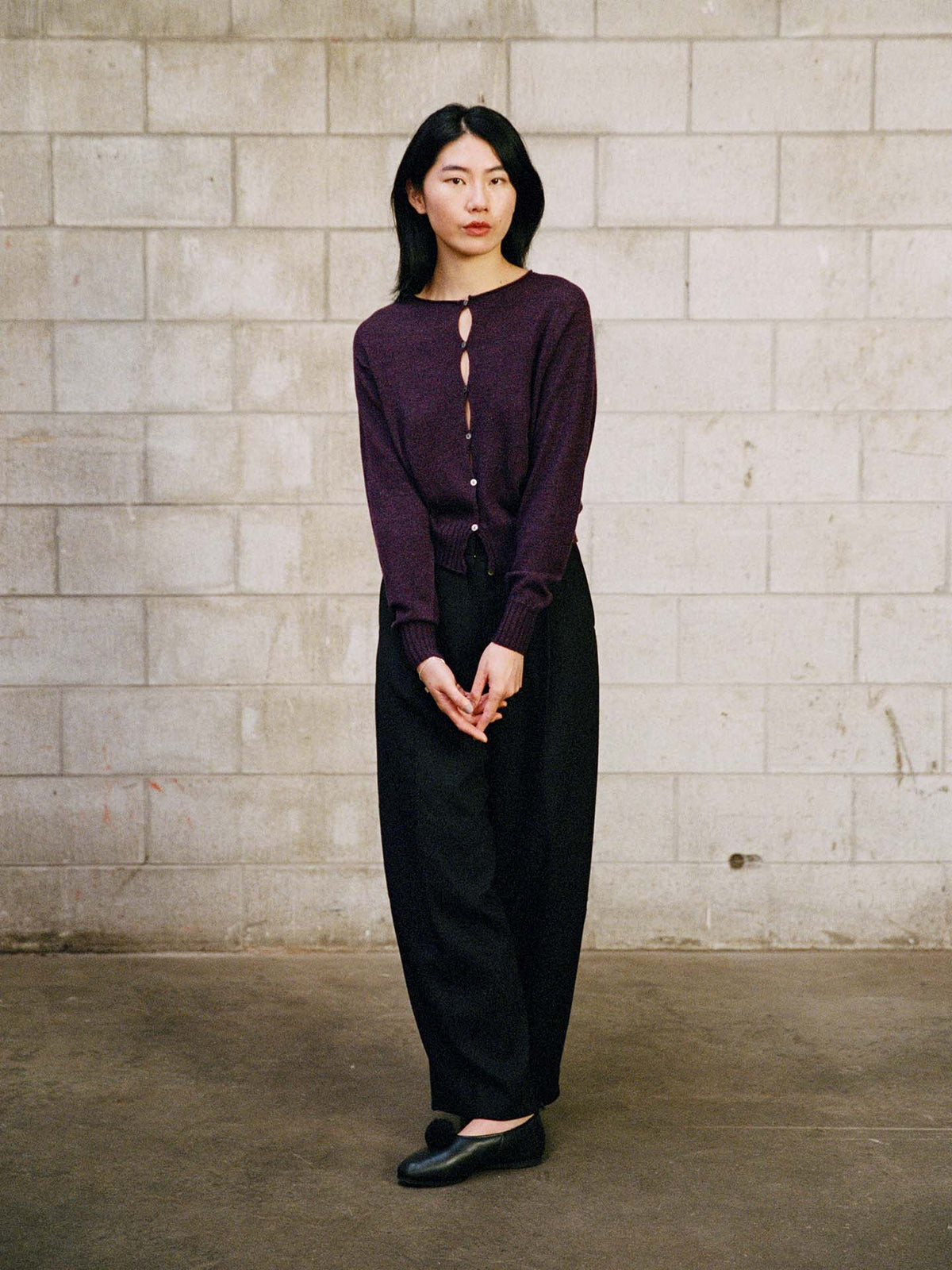 A person stands indoors against a gray cinder block wall, wearing the Anni Cardigan – Malbec in a relaxed fit, paired with black pants and shoes, hands clasped in front.