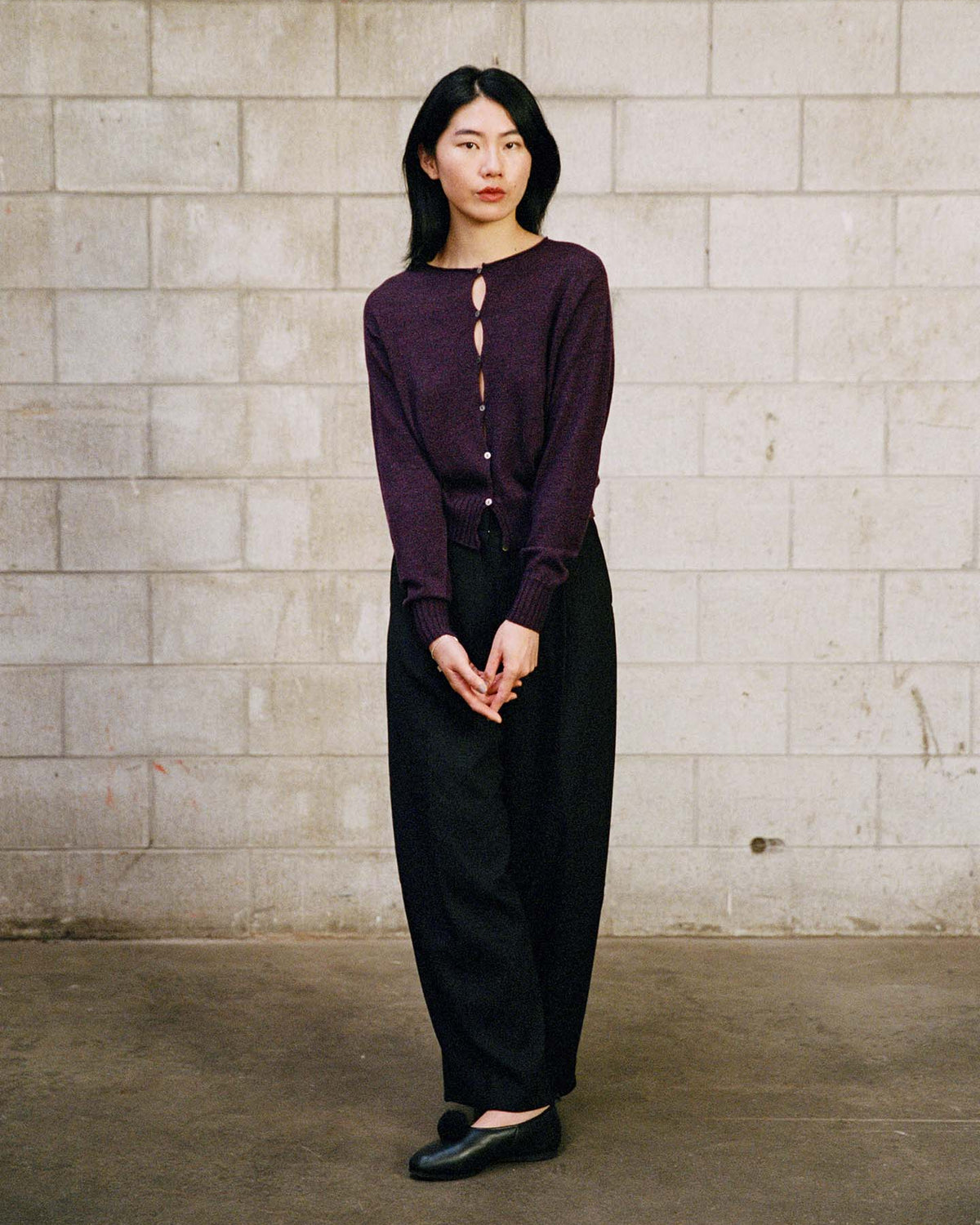 A person stands indoors against a gray cinder block wall, wearing the Anni Cardigan – Malbec in a relaxed fit, paired with black pants and shoes, hands clasped in front.