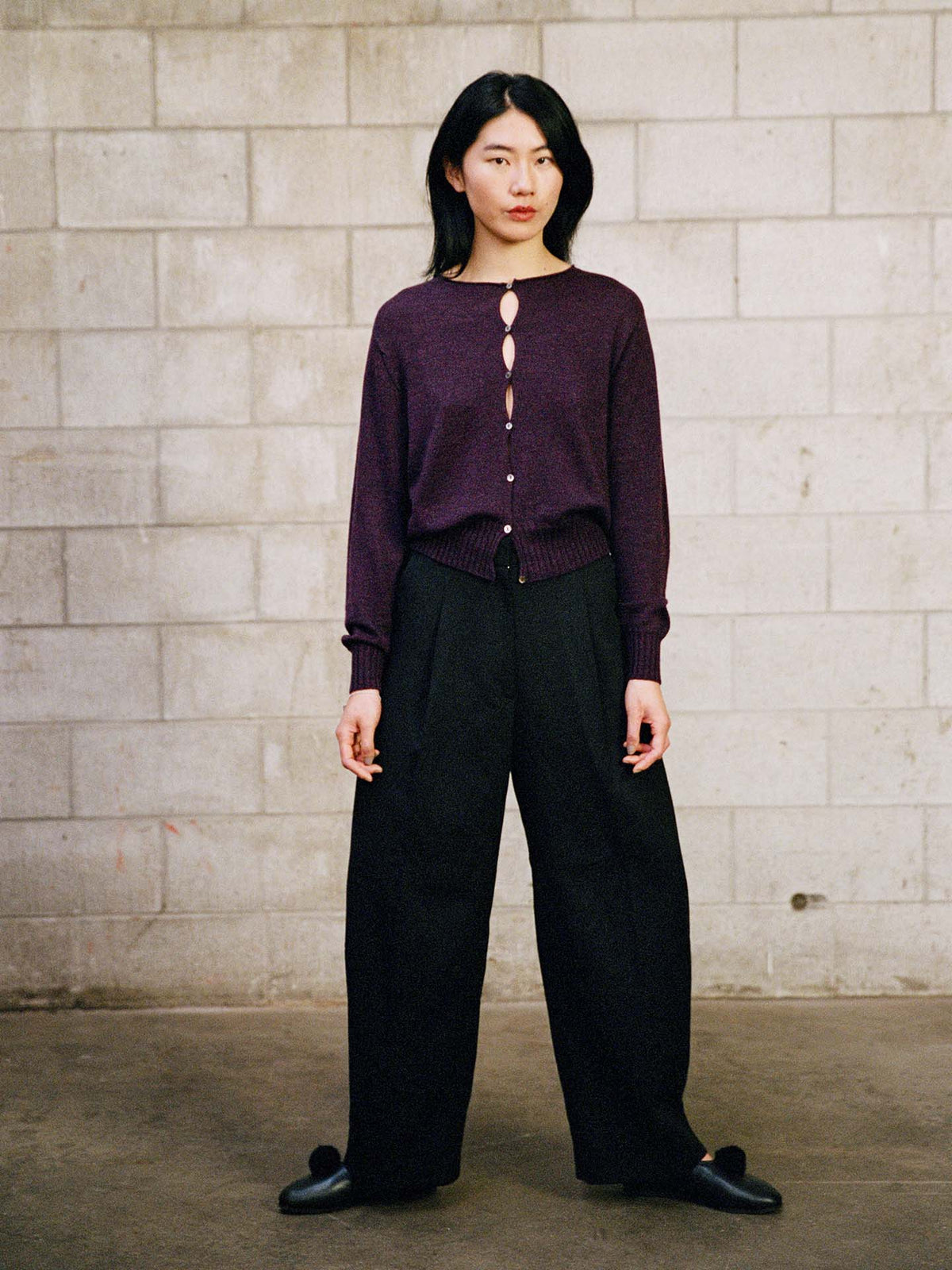 A person stands against a gray cinder block wall, wearing the Anni Cardigan – Malbec, black wide-leg pants, and black shoes.