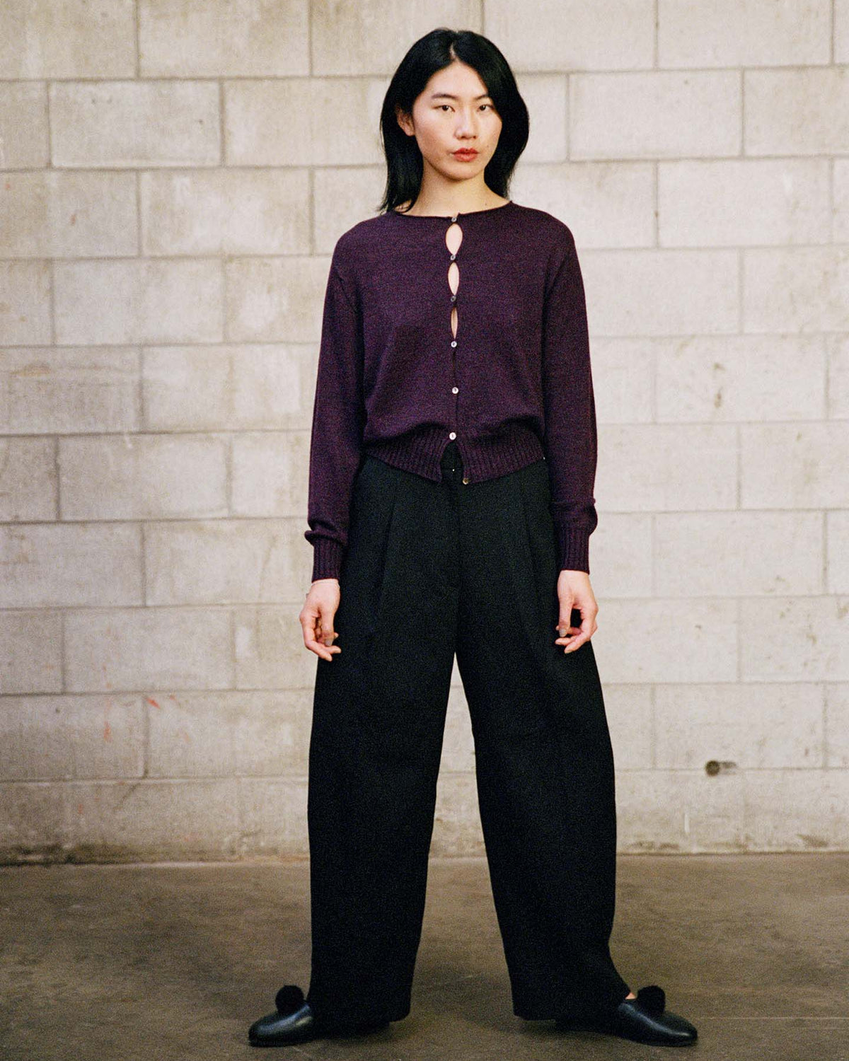 A person stands against a gray cinder block wall, wearing the Anni Cardigan – Malbec, black wide-leg pants, and black shoes.