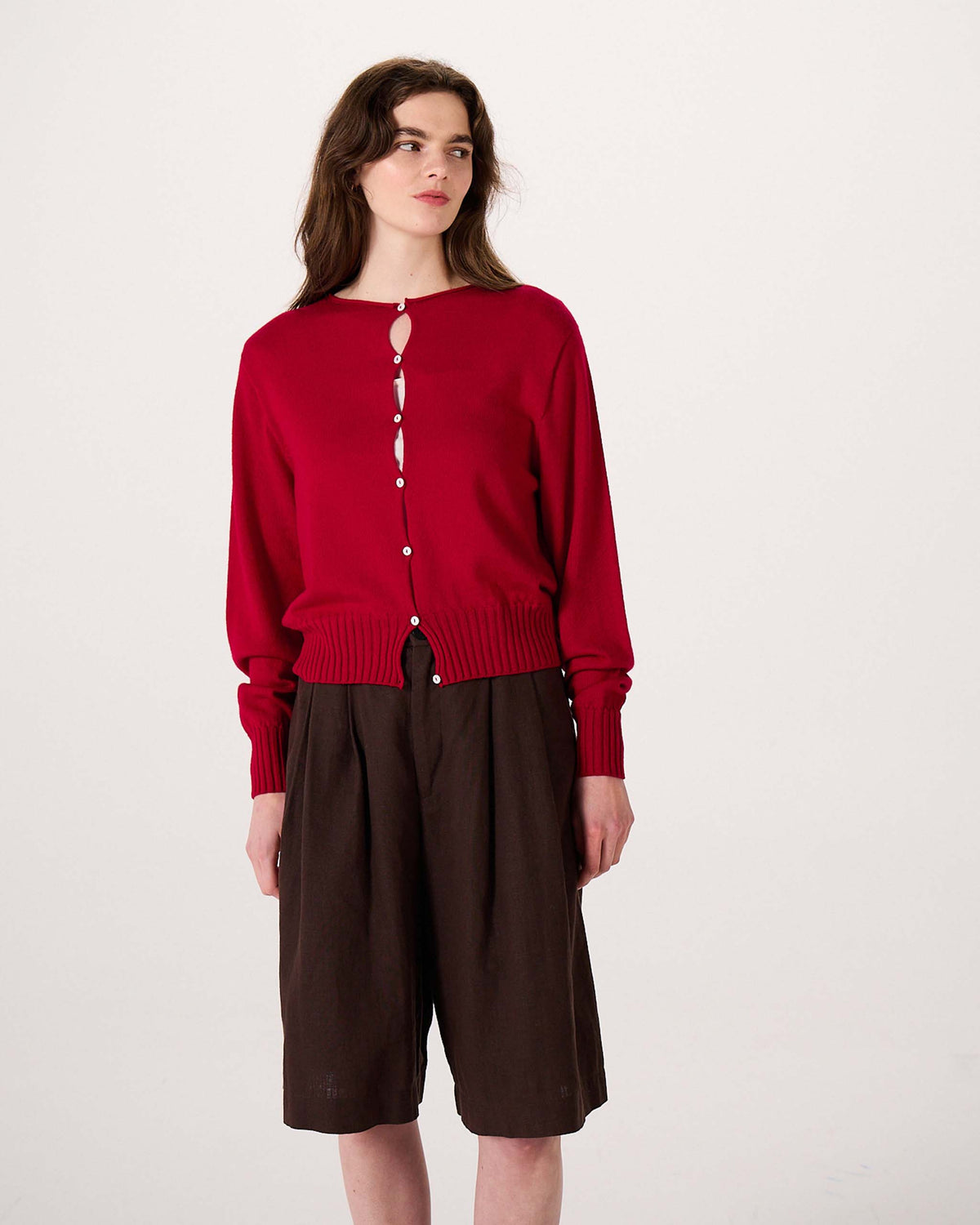 A person stands against a plain background wearing a red button-up cardigan and brown knee-length shorts, looking slightly to the side.