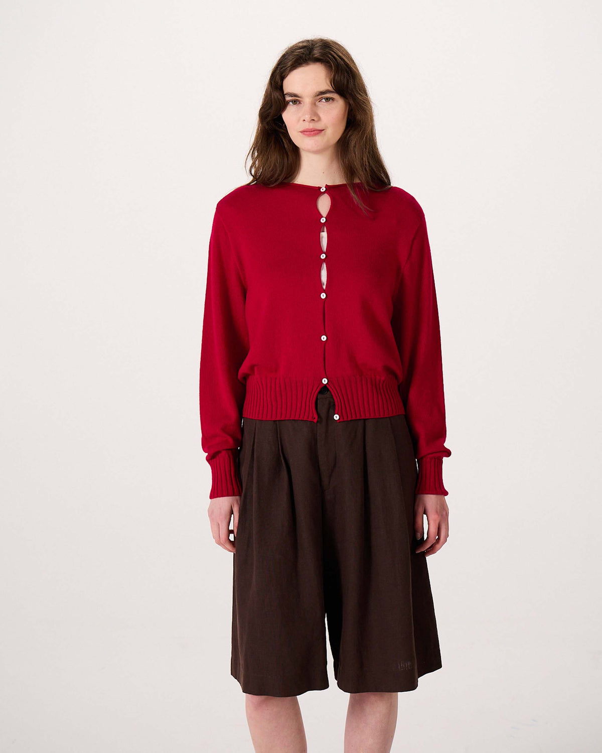 Woman standing against a plain background, wearing a red button-up cardigan with keyhole openings and dark brown knee-length shorts.