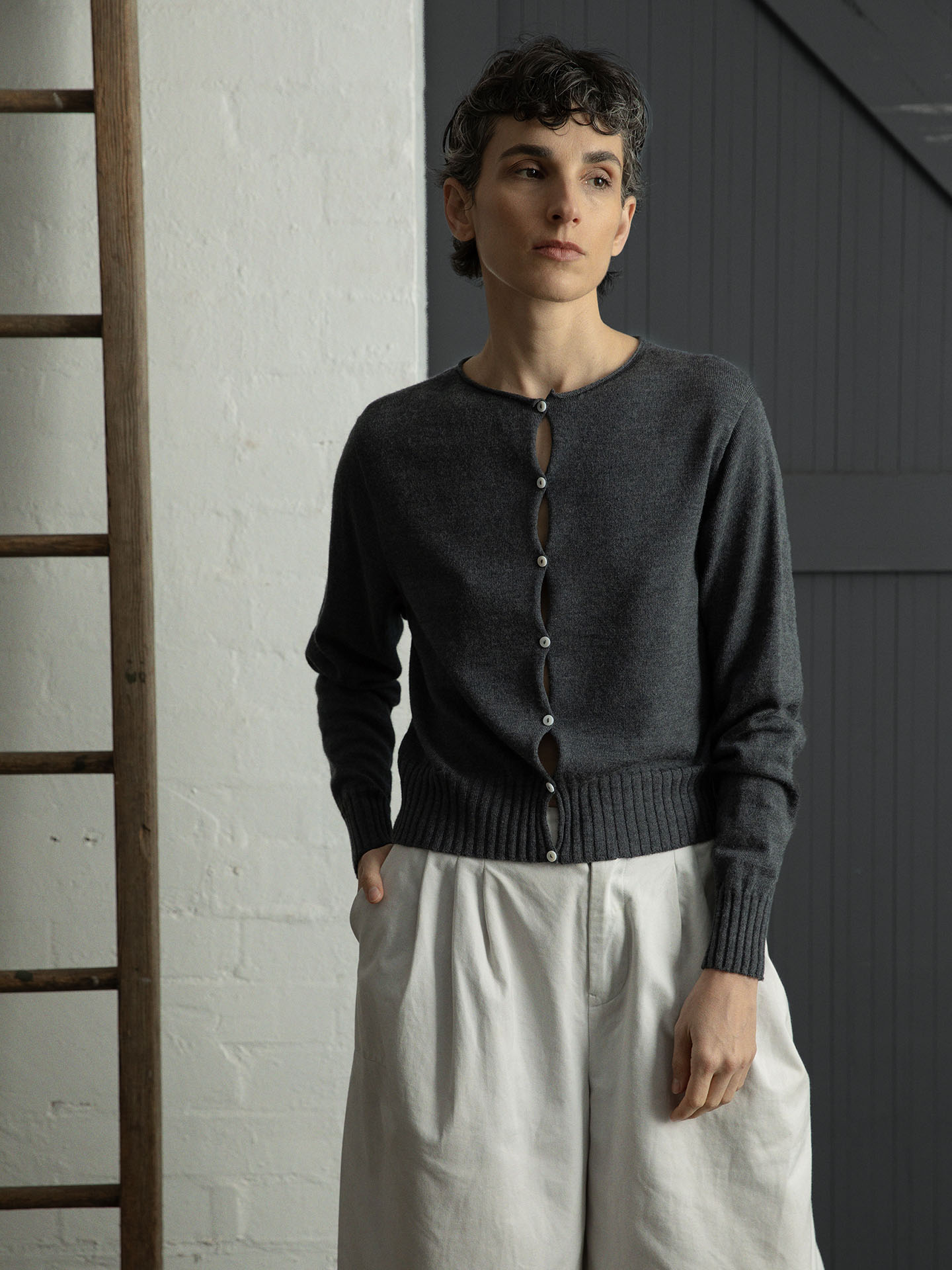 A person in cropped style wears the Anni Cardigan by Francie in Shadow Grey with light pants, standing against a white brick and gray paneled wall beside a wooden ladder.