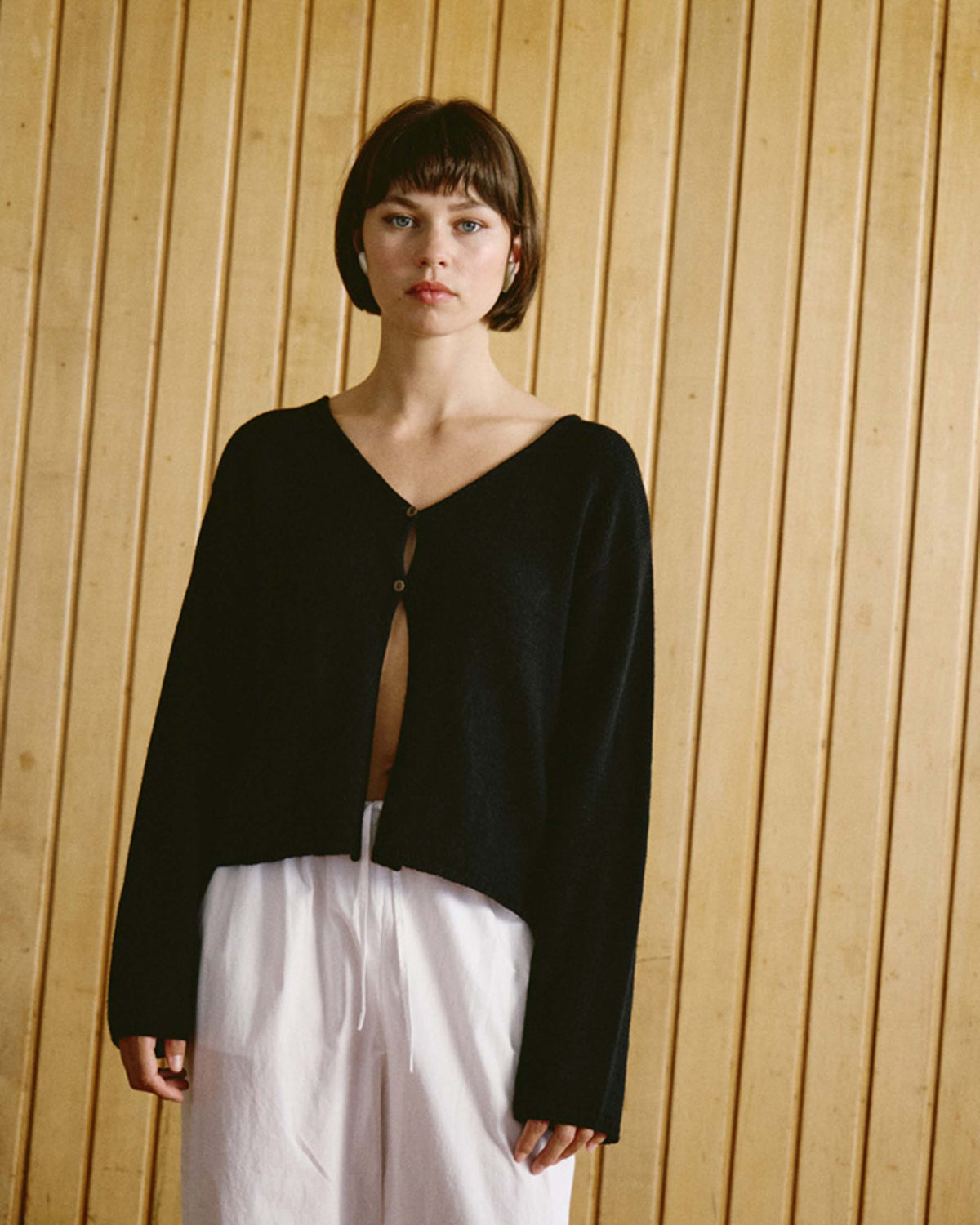 A person stands in front of a wooden panel wall, wearing a black long-sleeve top with one button fastened and loose white pants.