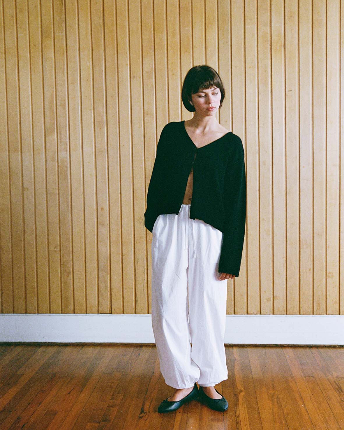 Person stands indoors on a wooden floor, wearing a black long-sleeve top, white loose pants, and black shoes, against a vertical wood panel wall.
