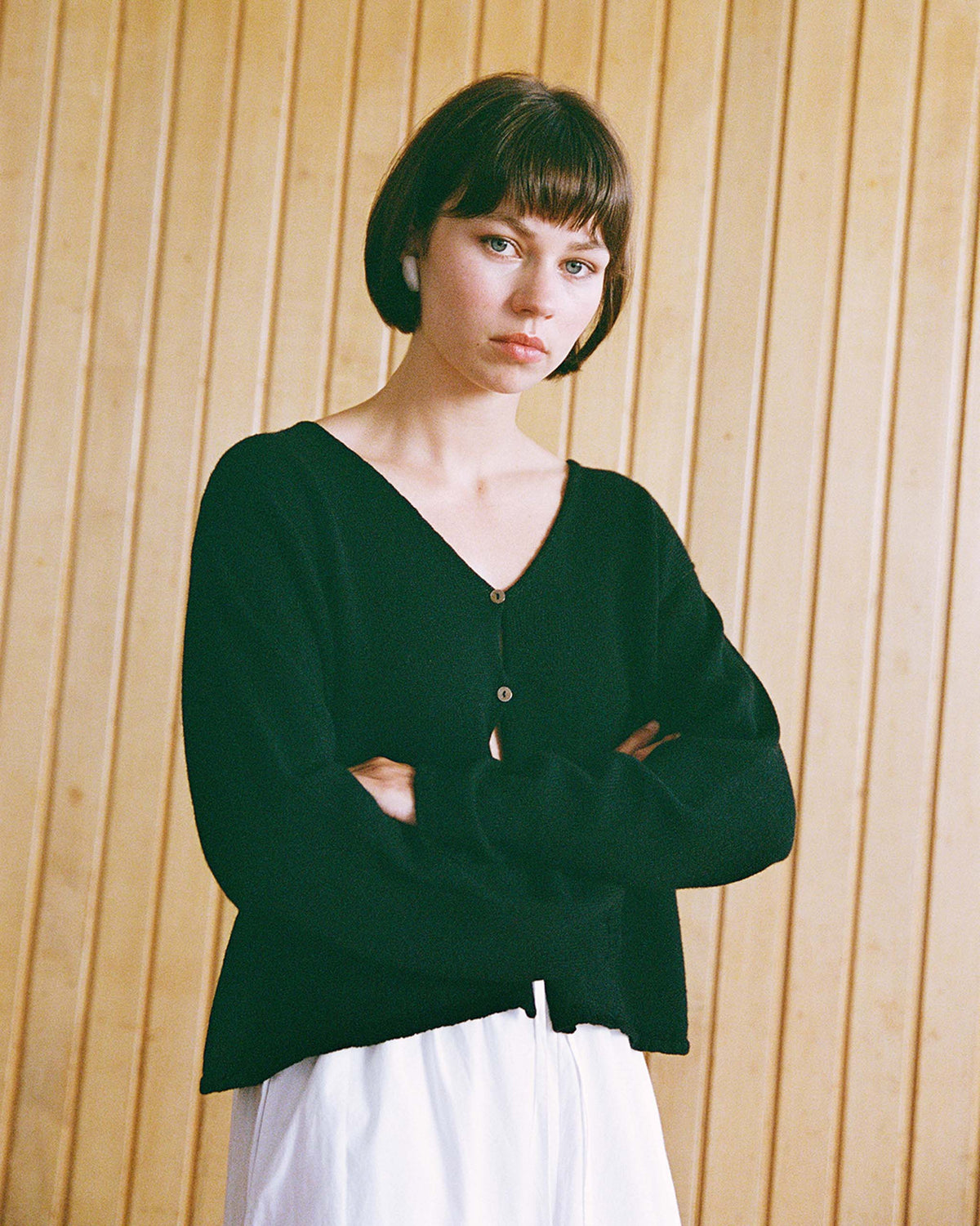 Person with short brown hair wearing a black buttoned sweater and white skirt stands with arms crossed in front of a wooden panel wall.