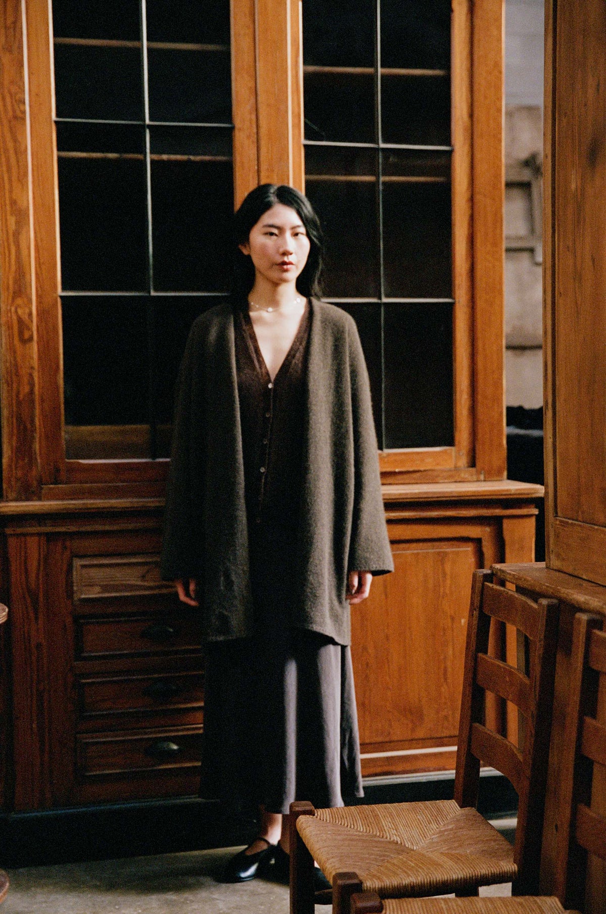 A woman stands indoors before a wooden cabinet, wearing the Cocoon Coat – Truffle in an oversized fit over a dark dress and black shoes. Wooden chairs appear in the foreground.